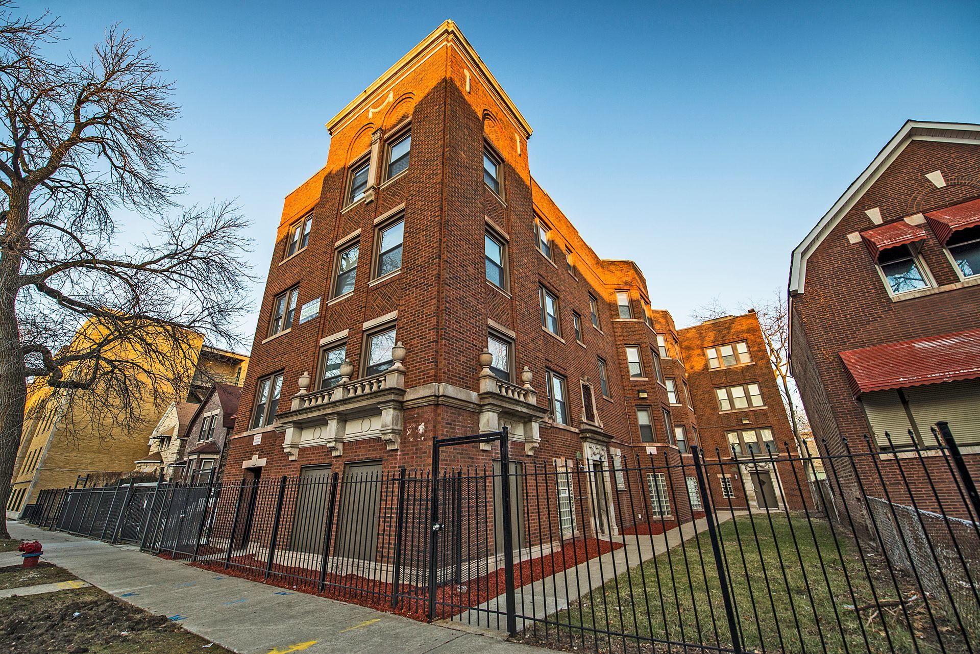 Brick apartment building with ornate details, metal fence, and sidewalk.
