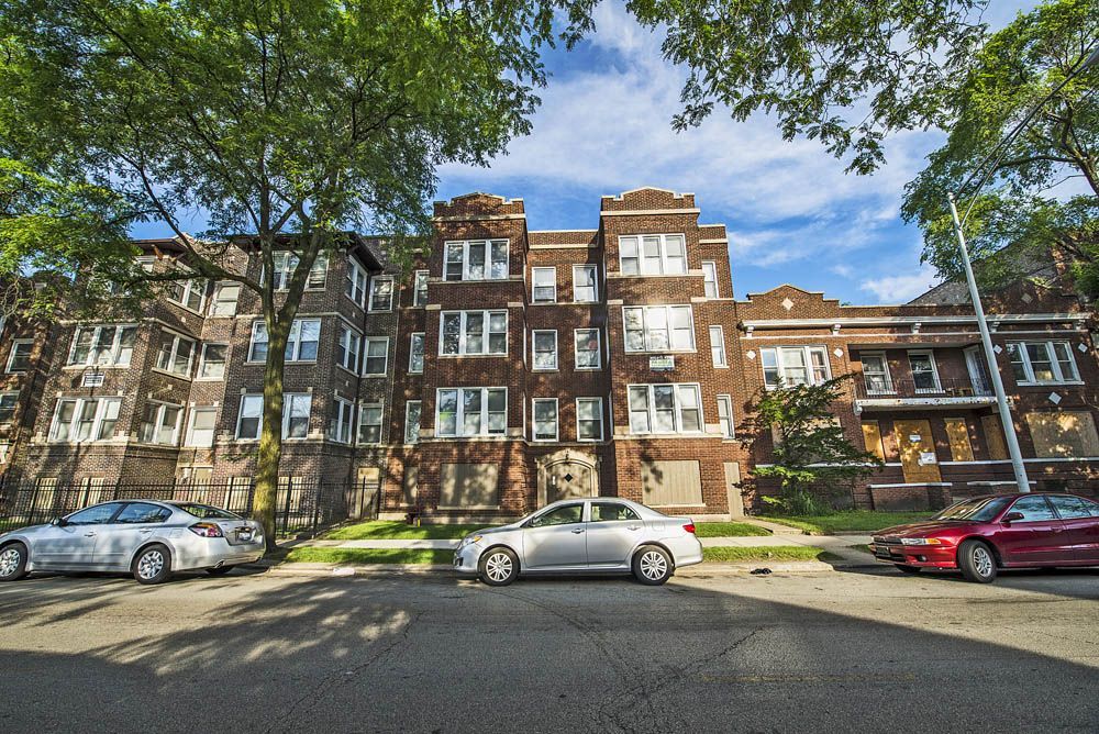 Three-story brick apartment building with cars parked on the street. Trees frame the building.