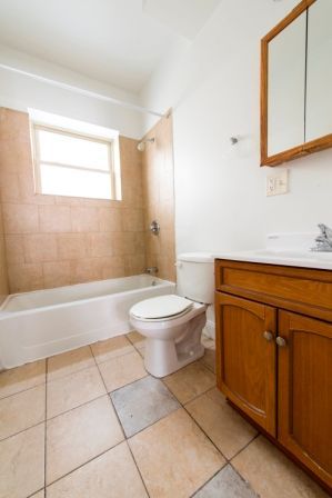 Bathroom with tan tiled walls, white tub and toilet, wooden vanity and medicine cabinet, window.