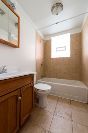 Bathroom with a white toilet, vanity, and bathtub, tan tile, and a window.