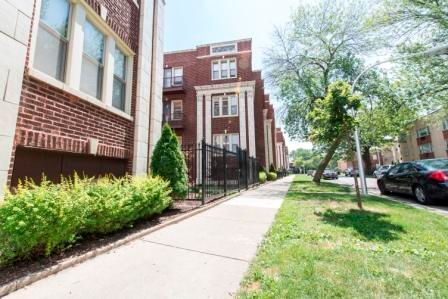 Brick apartment buildings with a sidewalk, a fence, and parked cars on a sunny day.