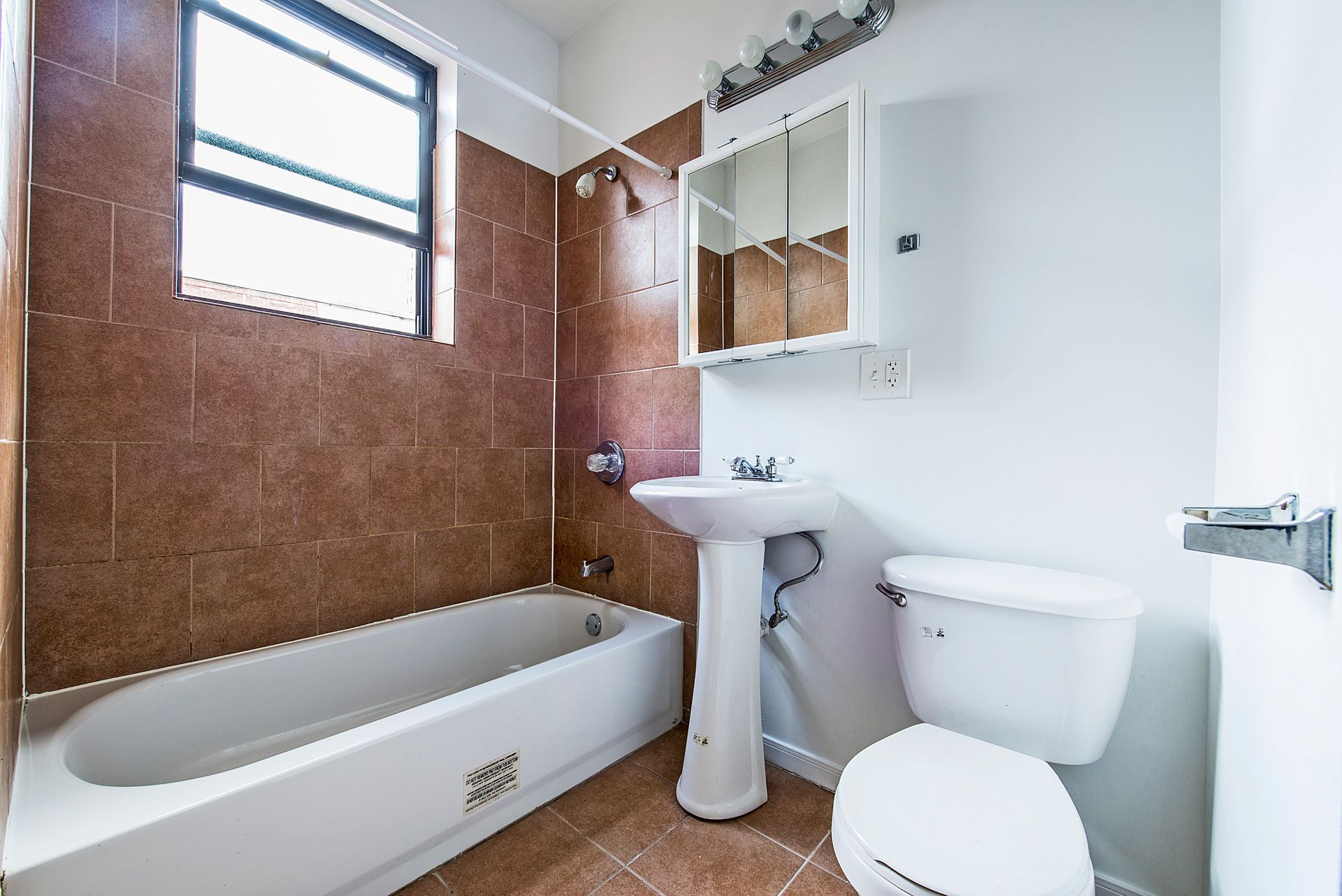 Bathroom with brown tiled walls, white fixtures: tub, toilet, pedestal sink, and medicine cabinet.