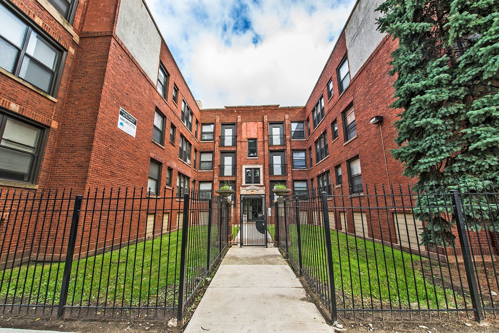 Red brick apartment building with a pathway leading to the entrance, fenced in with black wrought iron.