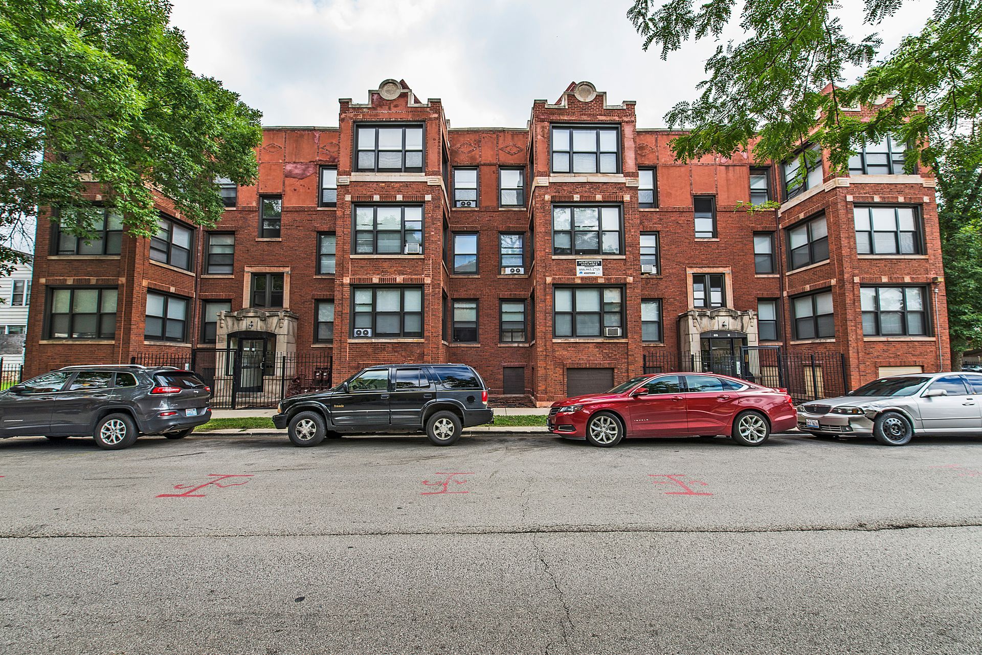 Brick apartment building with parked cars on the street. Green trees frame the sides.