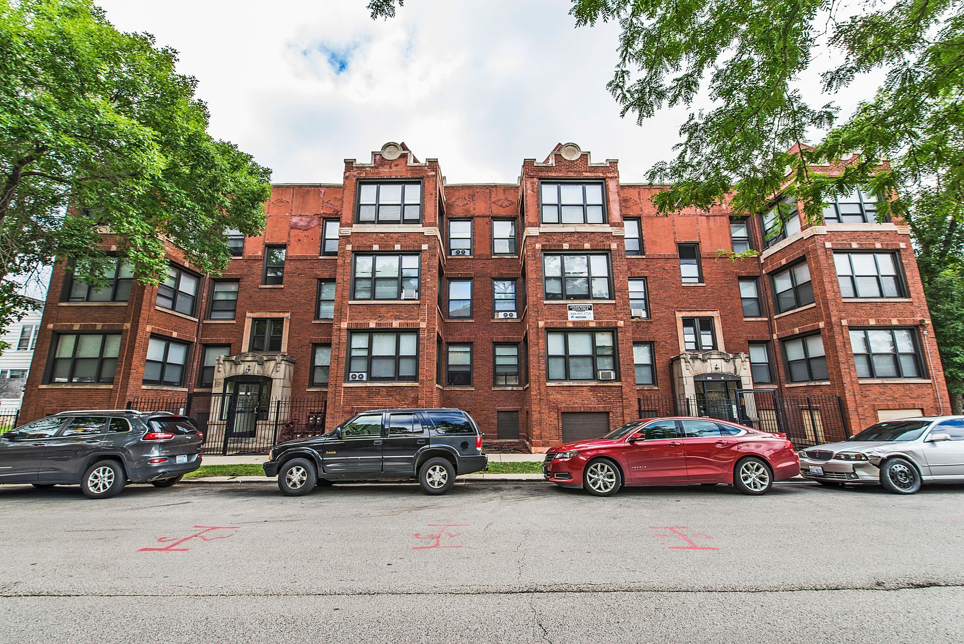 Red brick apartment building with parked cars on a tree-lined street under a cloudy sky.