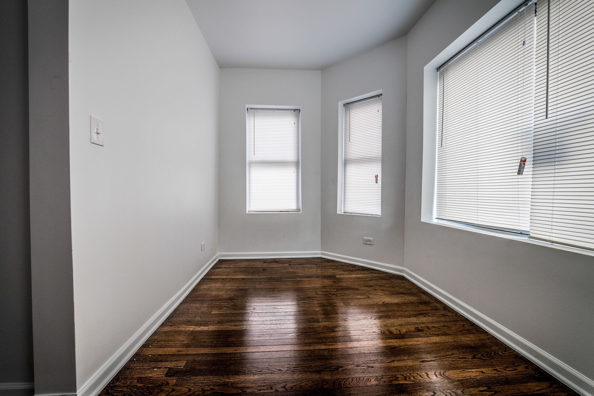 Empty room with wooden floor, white walls, and three windows with blinds.