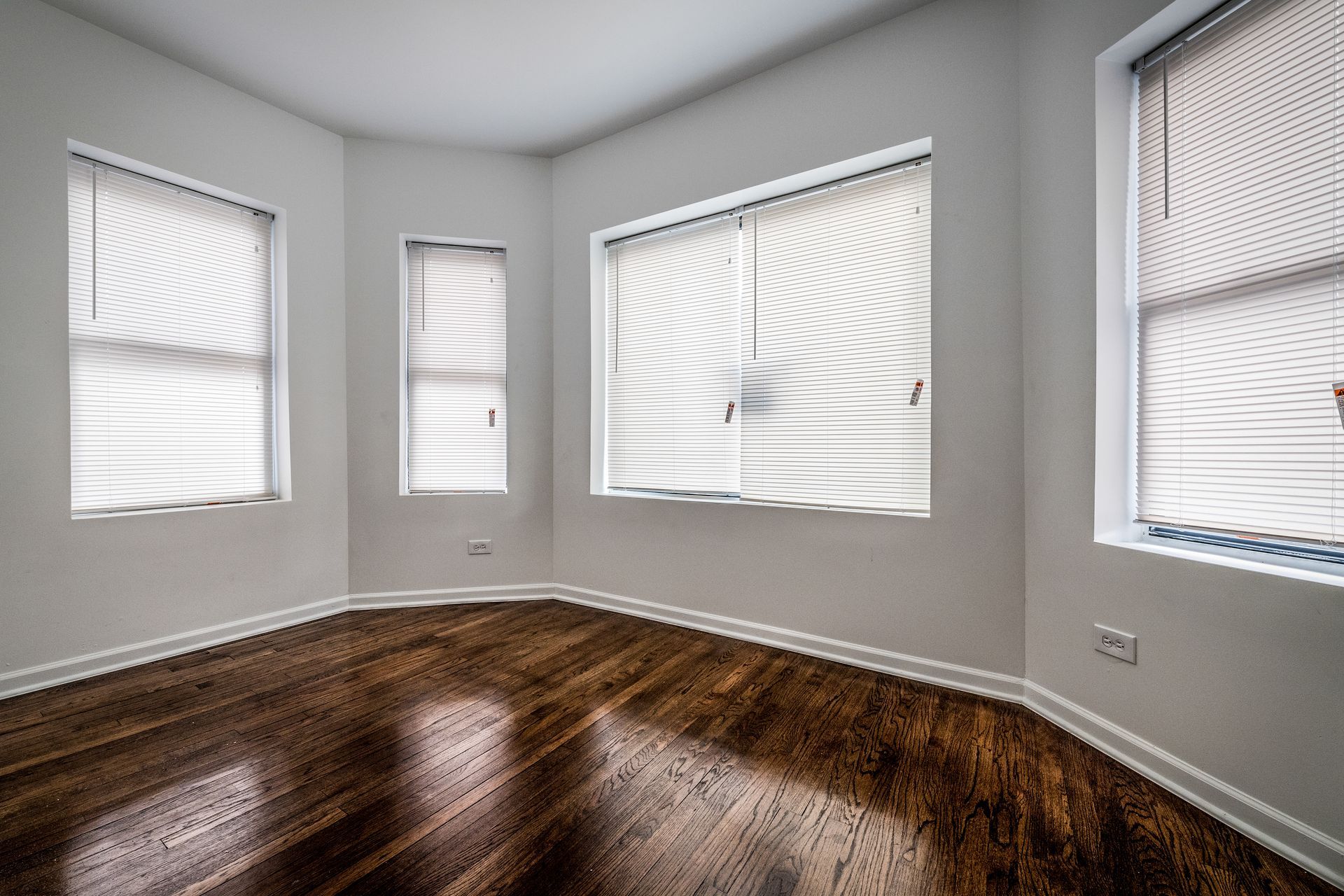 Empty room with wood floor, white walls, and blinds on windows.