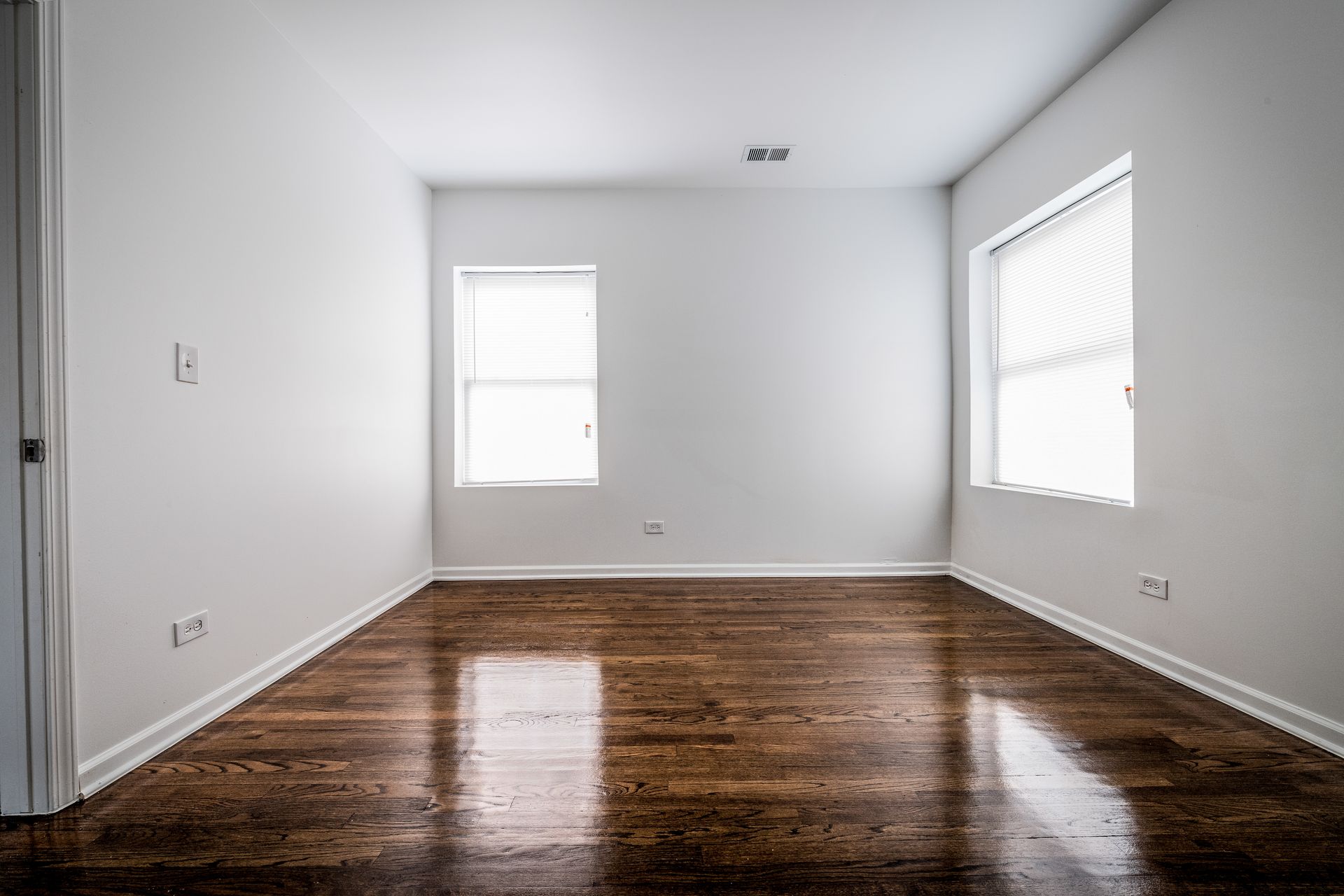 Empty room with hardwood floors, white walls, two windows, and closed blinds.