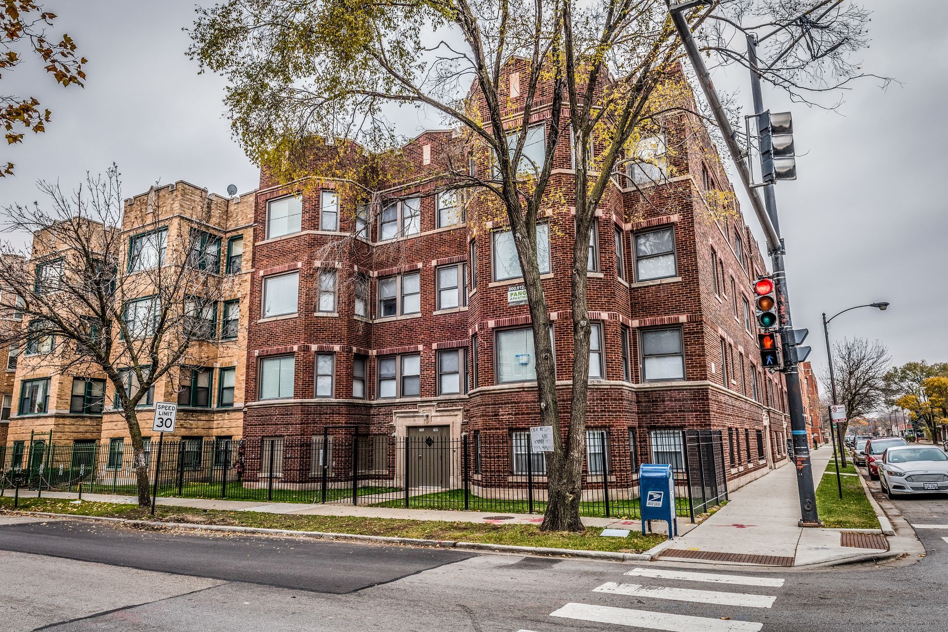 Brick apartment building on a street corner, with trees and a cloudy sky.