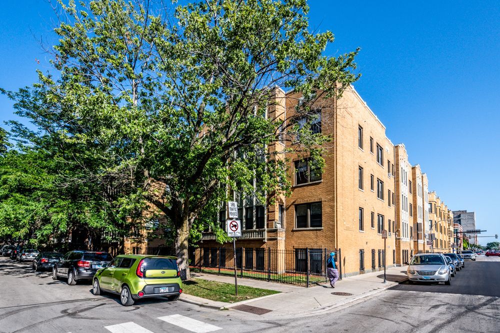 Apartment building on a sunny street with parked cars and a pedestrian on the sidewalk.