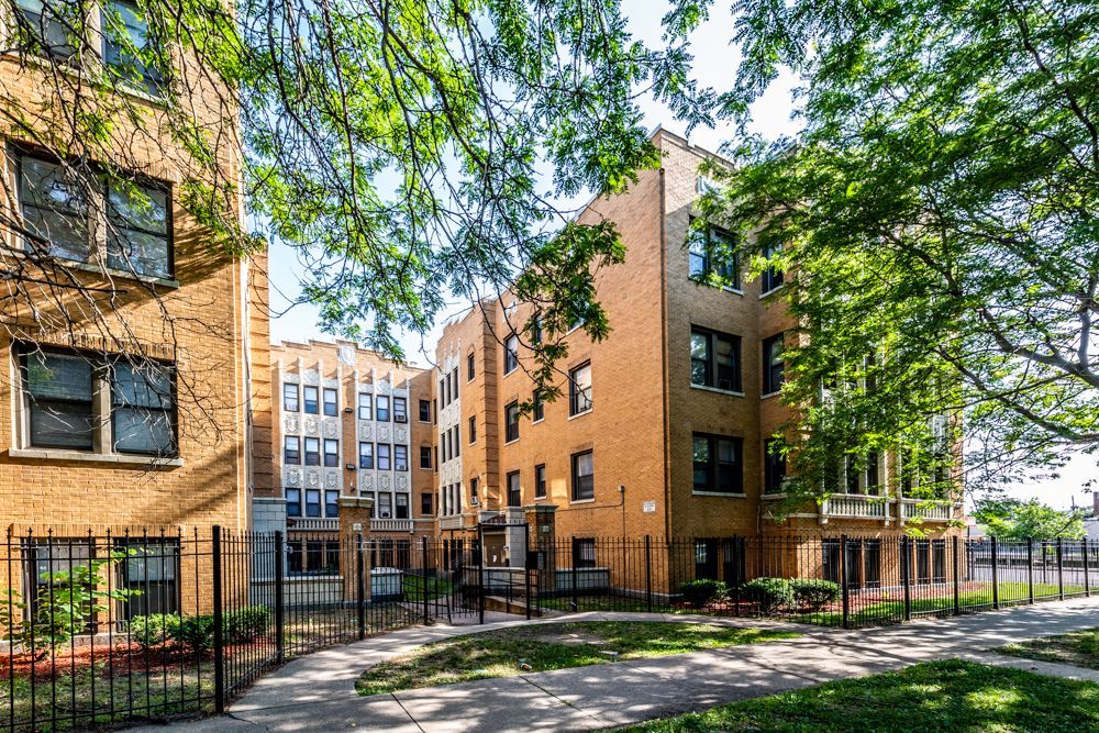 Brick apartment buildings with arched entryways, trees, and a wrought-iron fence.