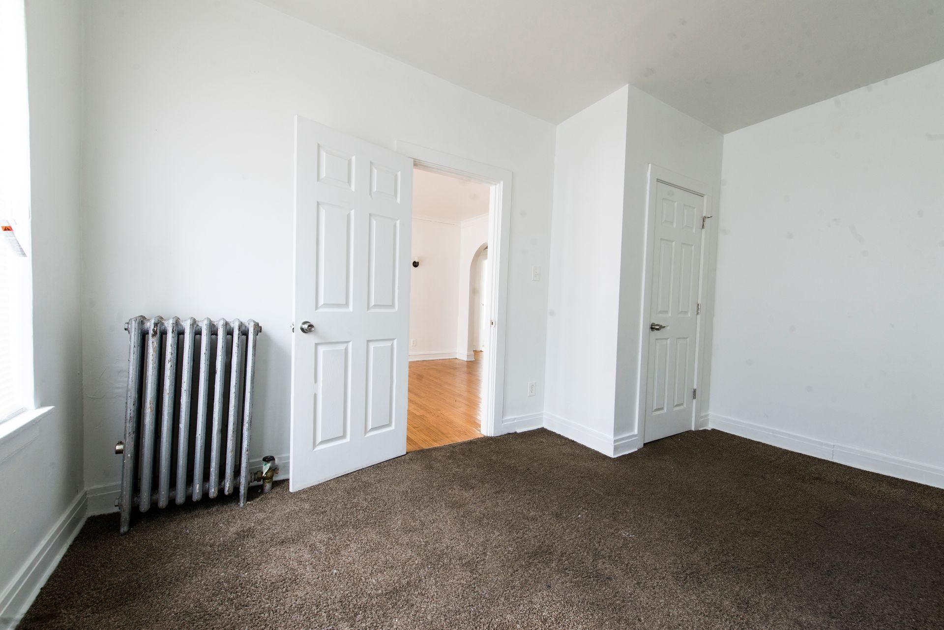 Empty room with brown carpet, white walls, two white doors, and a radiator.