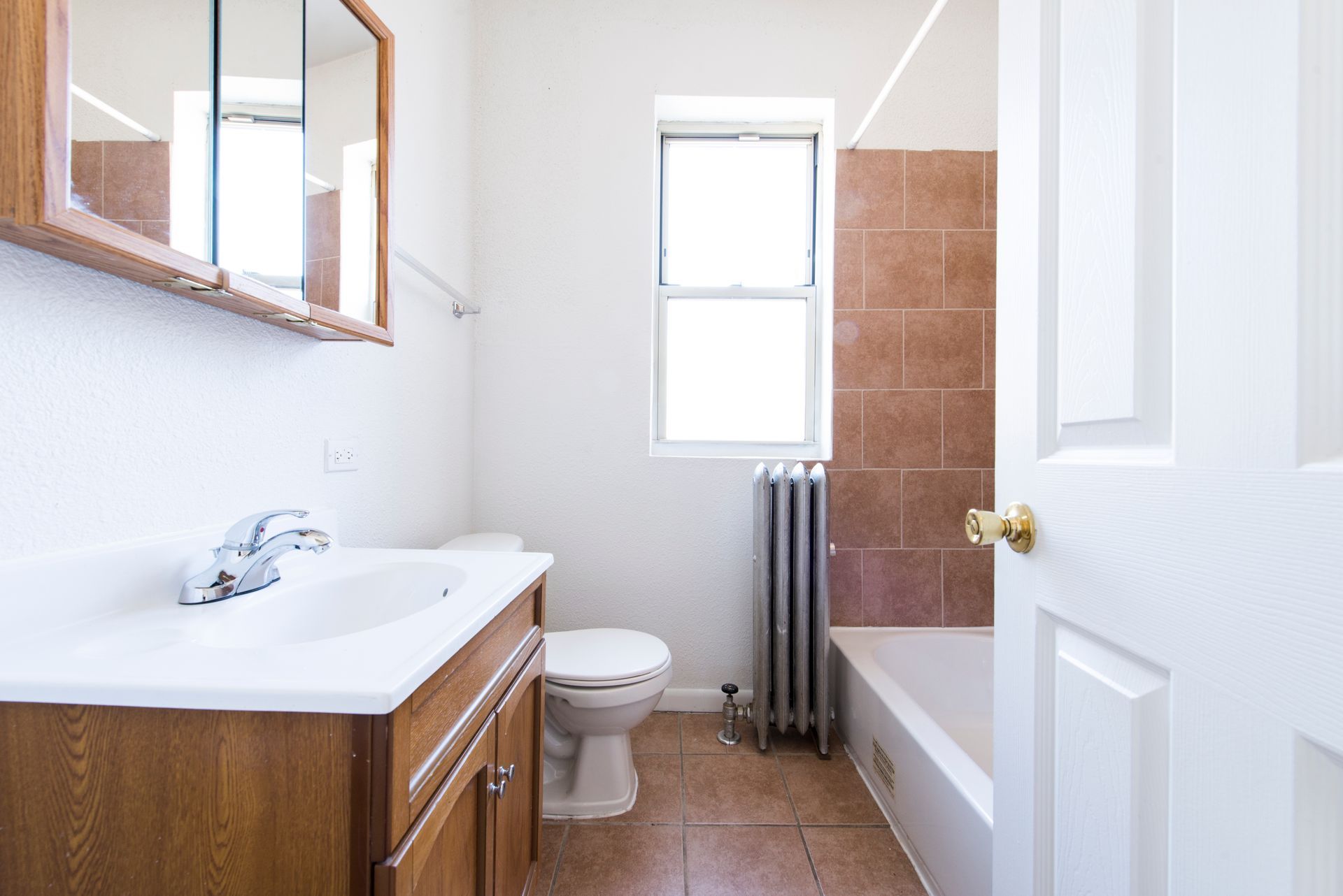 Bathroom with a white sink, toilet, and bathtub. Brown tile and cabinet. A window is open. The door is open.