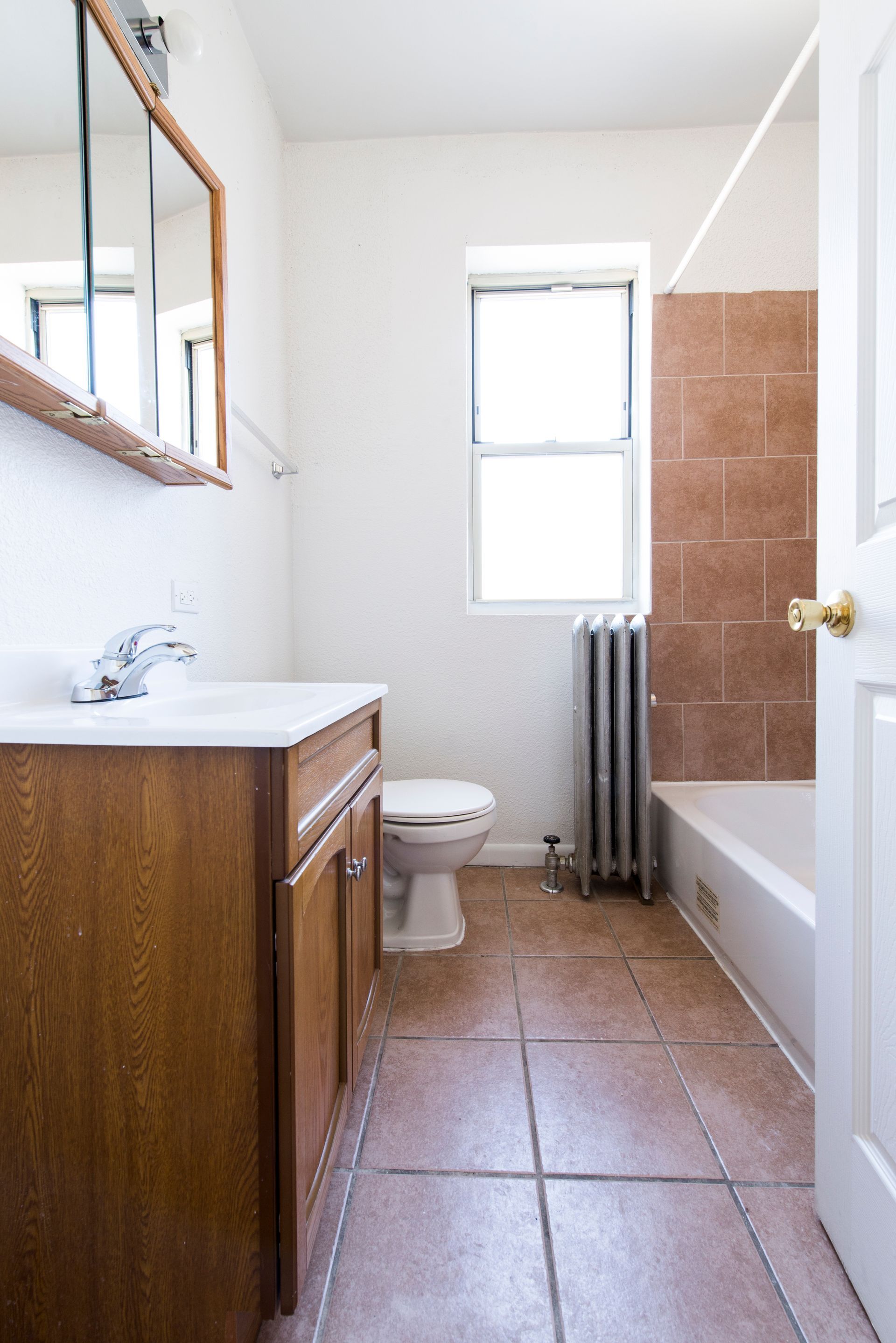 Bathroom with vanity, toilet, tub, and window; brown tile floor, white walls and door.