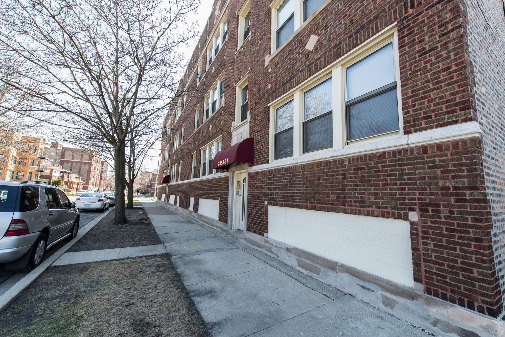Brick apartment building on a city street, silver car parked in front, sidewalk, bare tree.