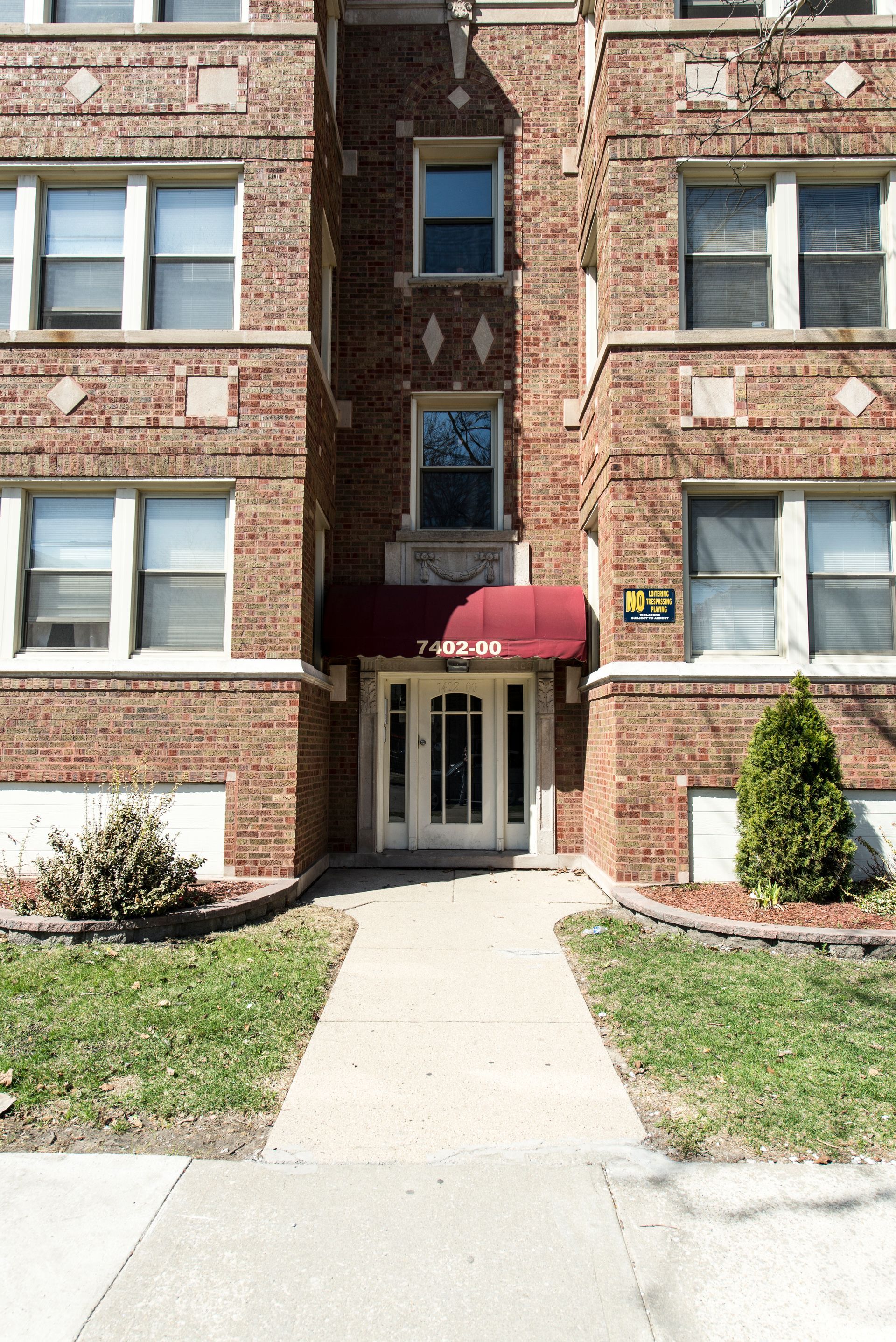 Brick apartment building with red awning over entryway and concrete path.