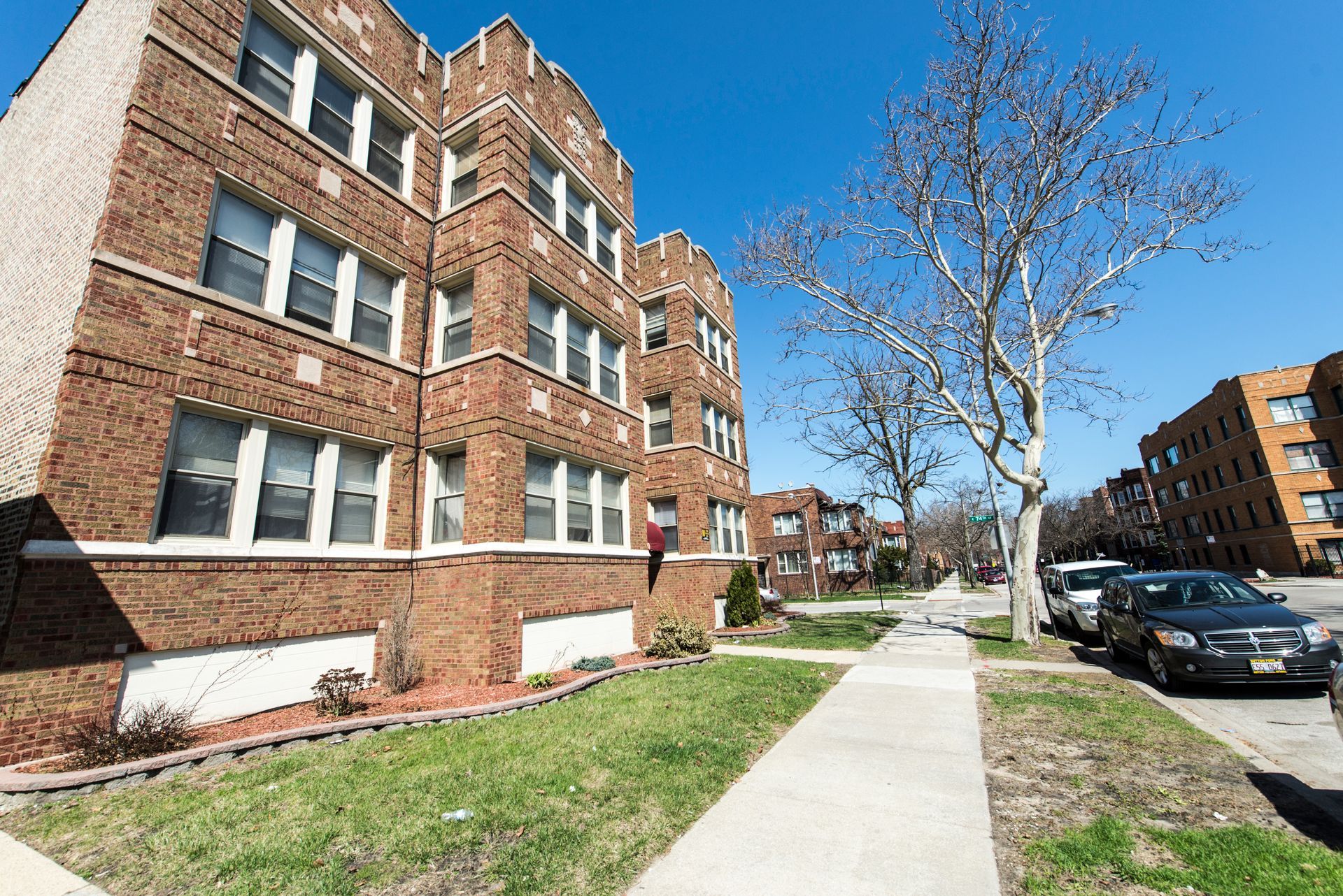 Brick apartment building on a sunny street with parked cars and a sidewalk.