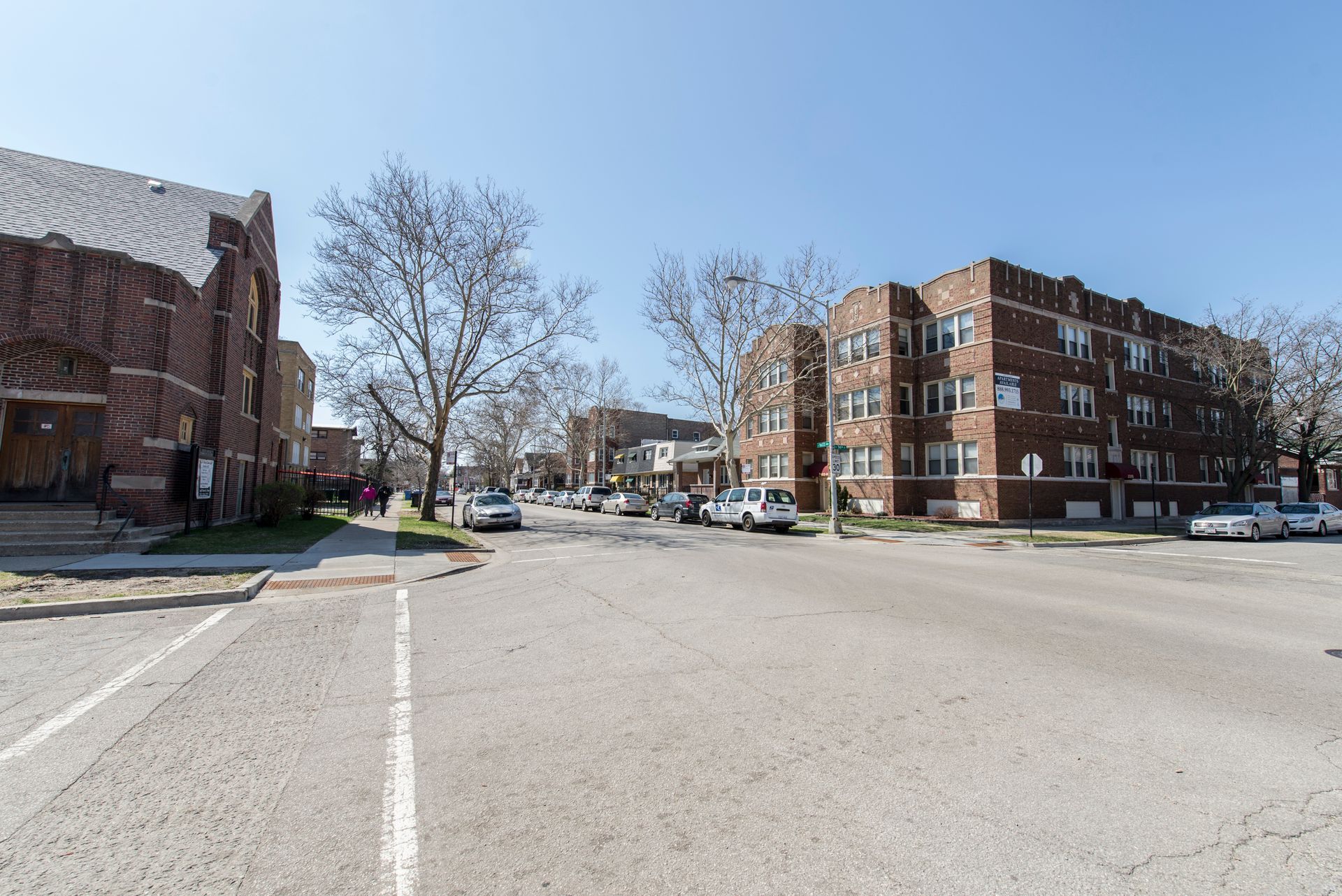 Street scene with brick buildings, trees, parked cars, and clear sky.