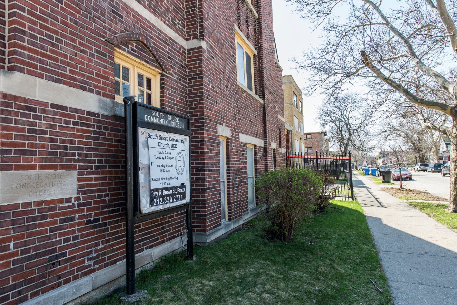 Brick building with a sign on the lawn; sidewalk, street, and trees are visible.