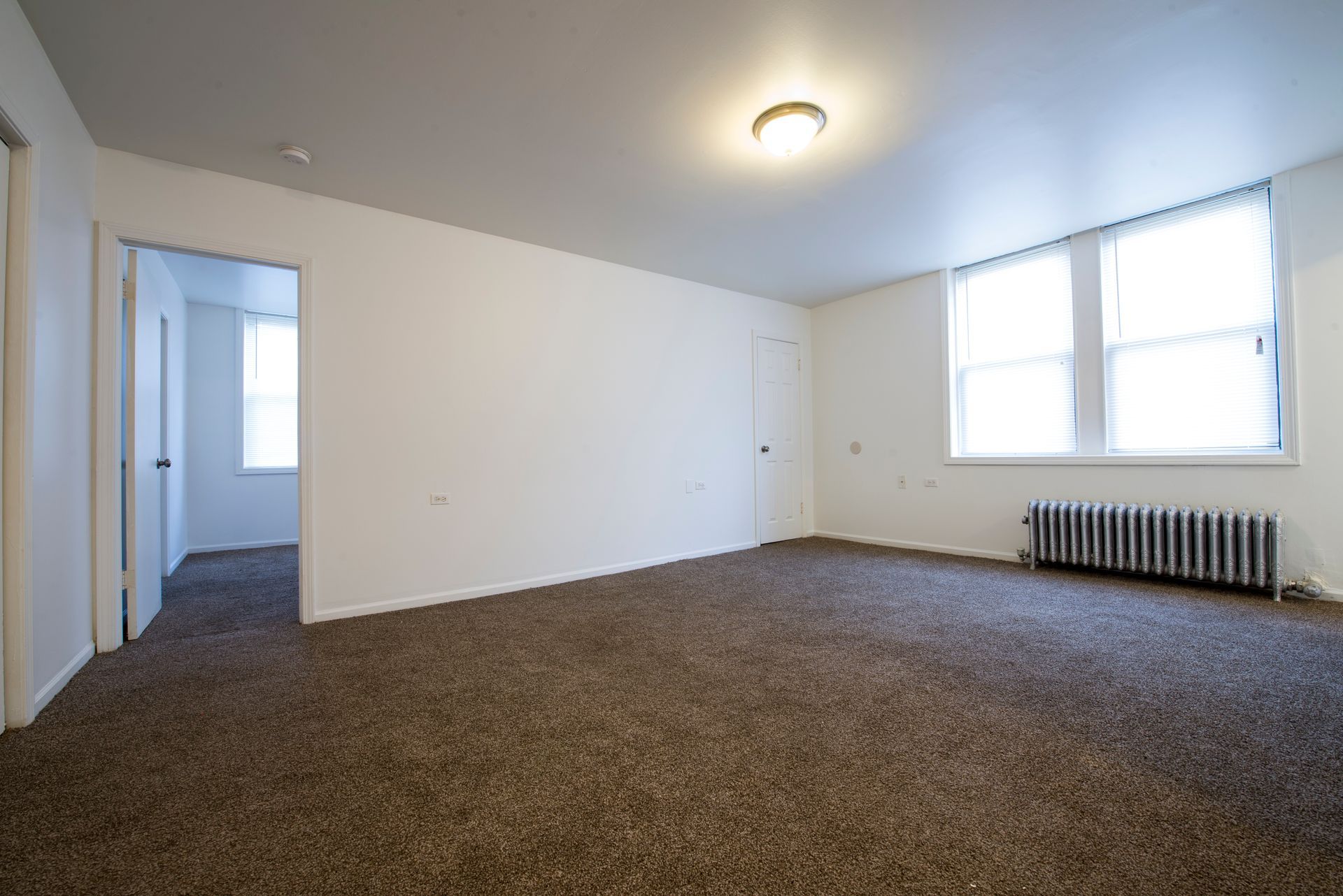 Empty apartment room with brown carpet, white walls, and two windows.