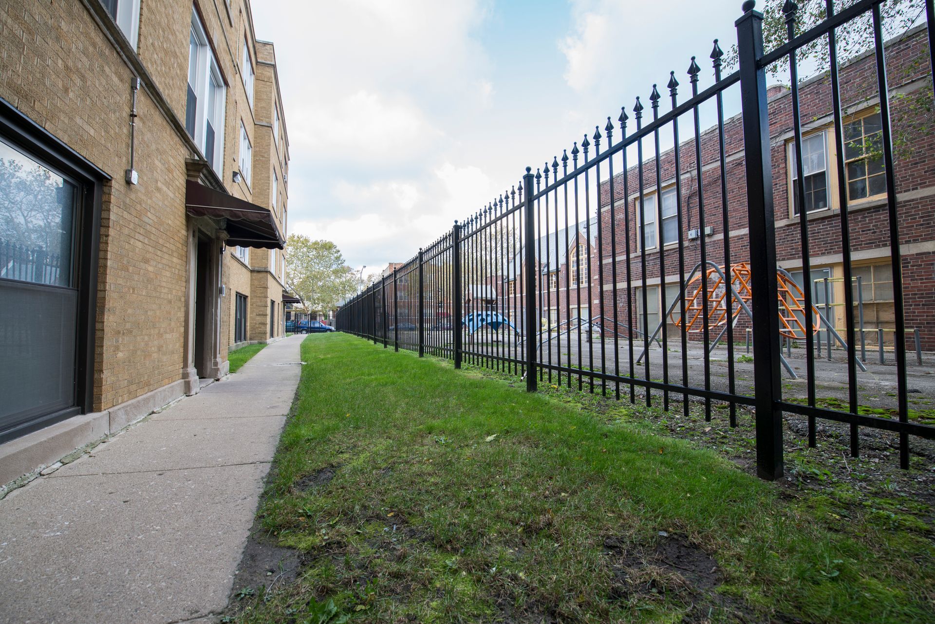 Low-angle view of a long, black metal fence beside a building and narrow grass strip.