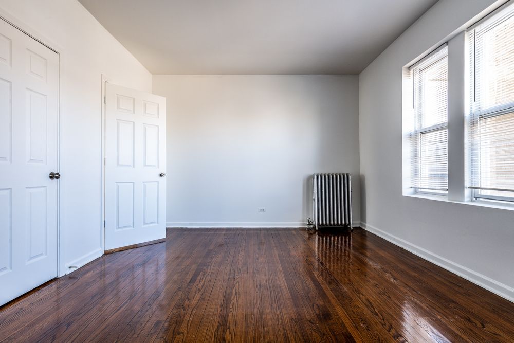 Empty room with hardwood floors, white walls, two doors, a radiator, and a window.
