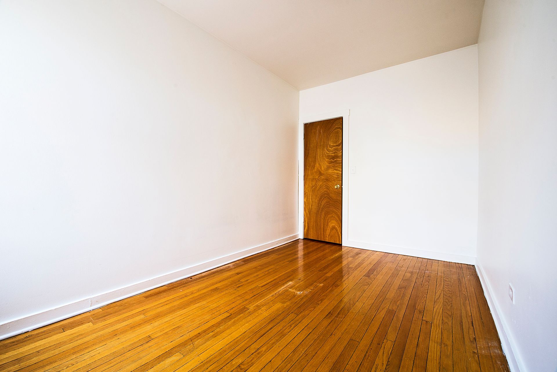 Empty room with wooden floor, white walls, and a closed brown door.