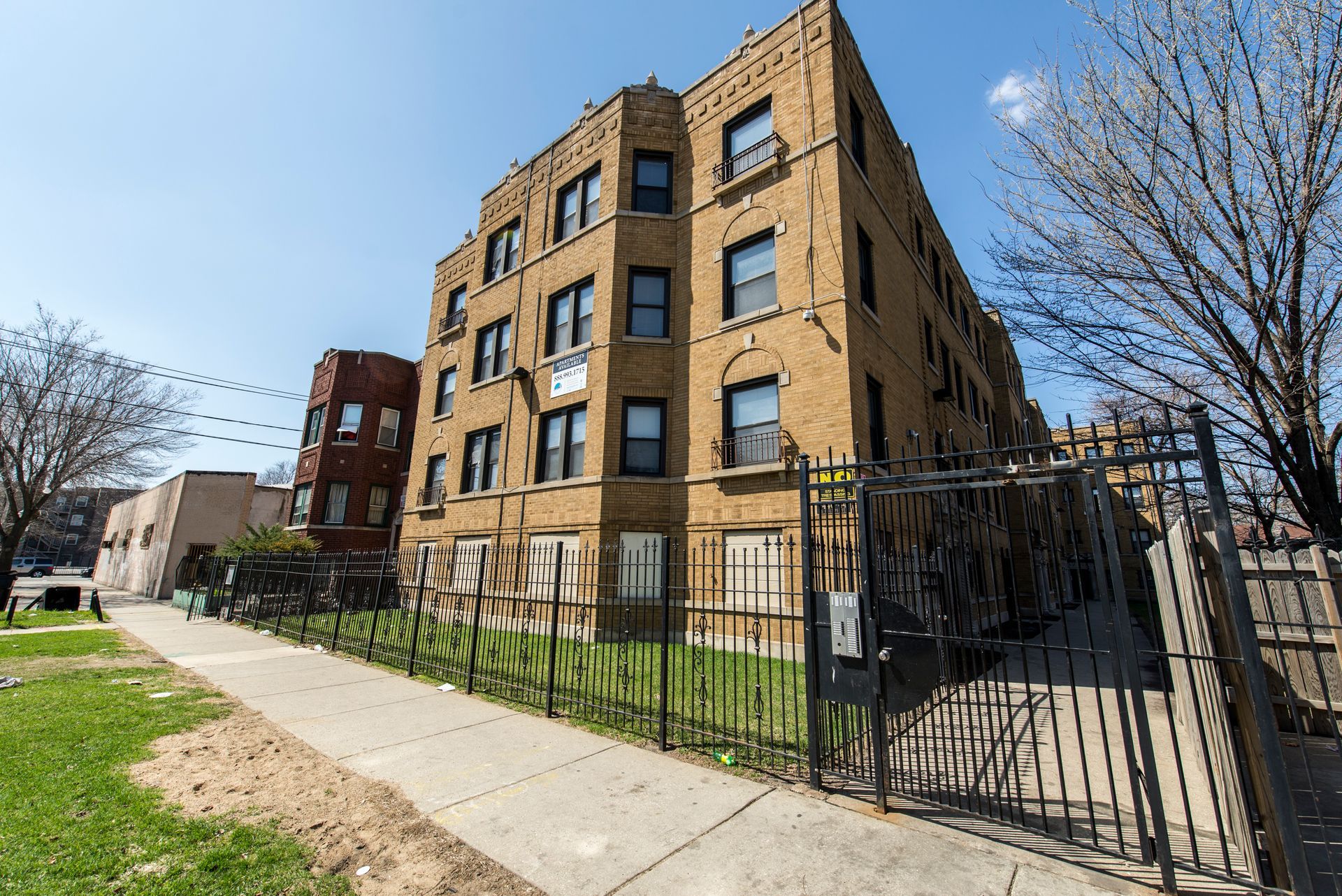 Brown brick apartment building behind a black metal fence on a sunny day.