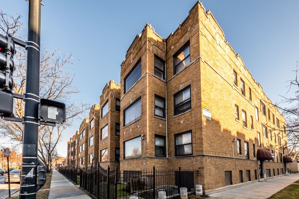 Brick apartment building on a sunny street, corner view with dark metal fence, blue sky.