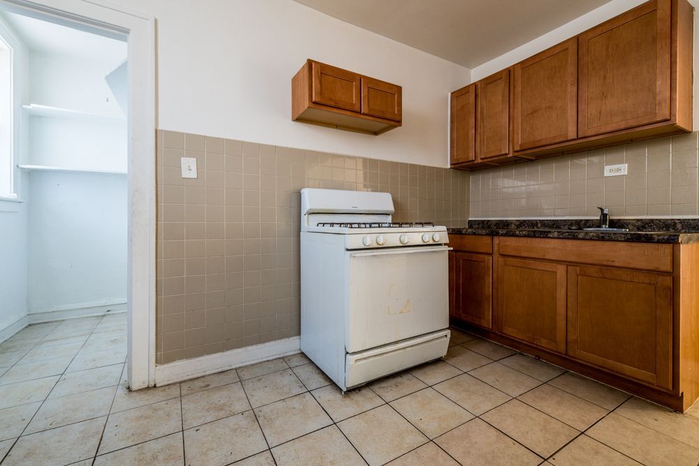 Kitchen with white stove, brown cabinets, and tiled walls and floor. A pantry is visible in the doorway.