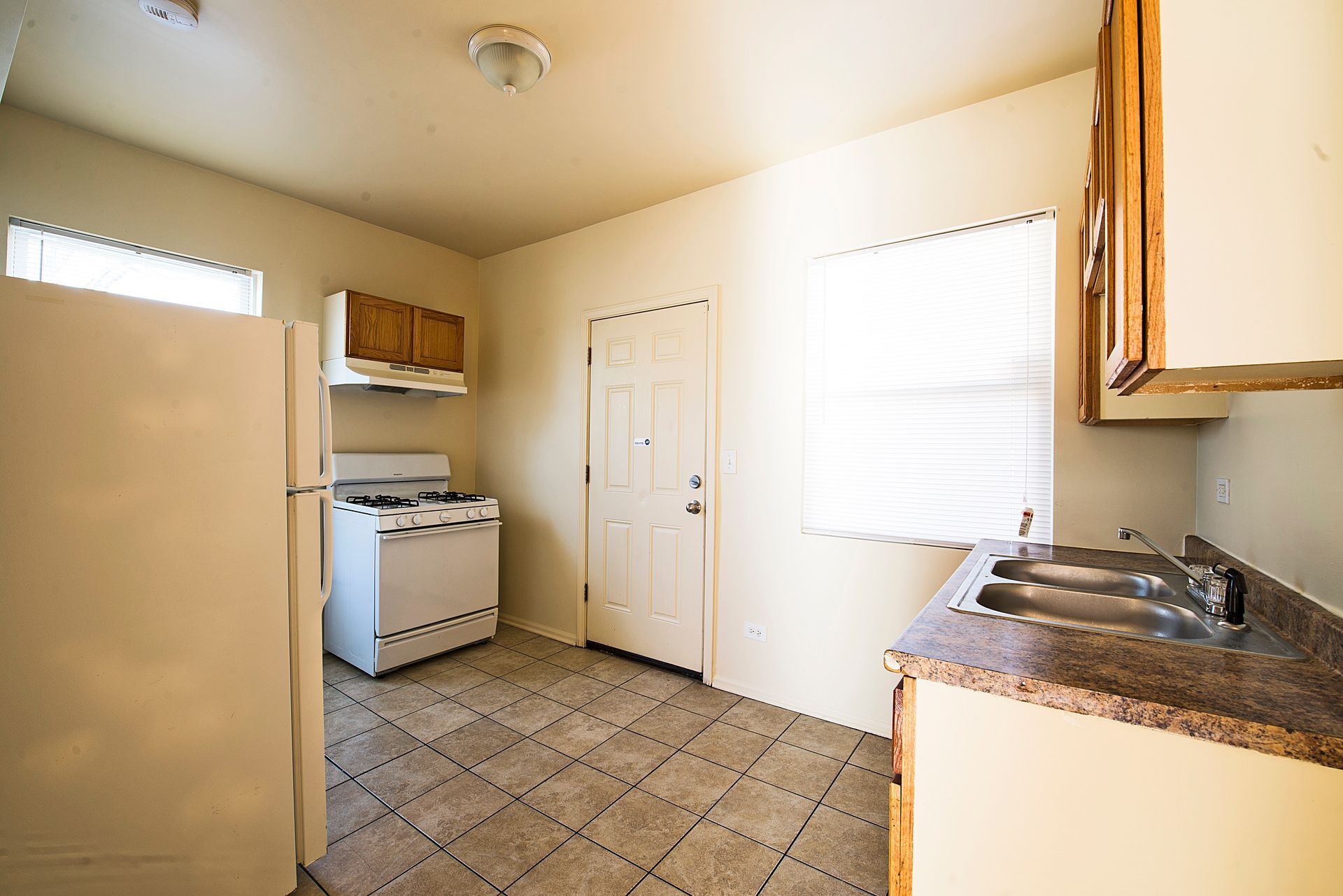 A small kitchen with a refrigerator, stove, cabinets, a sink, and a door. Tile floor.