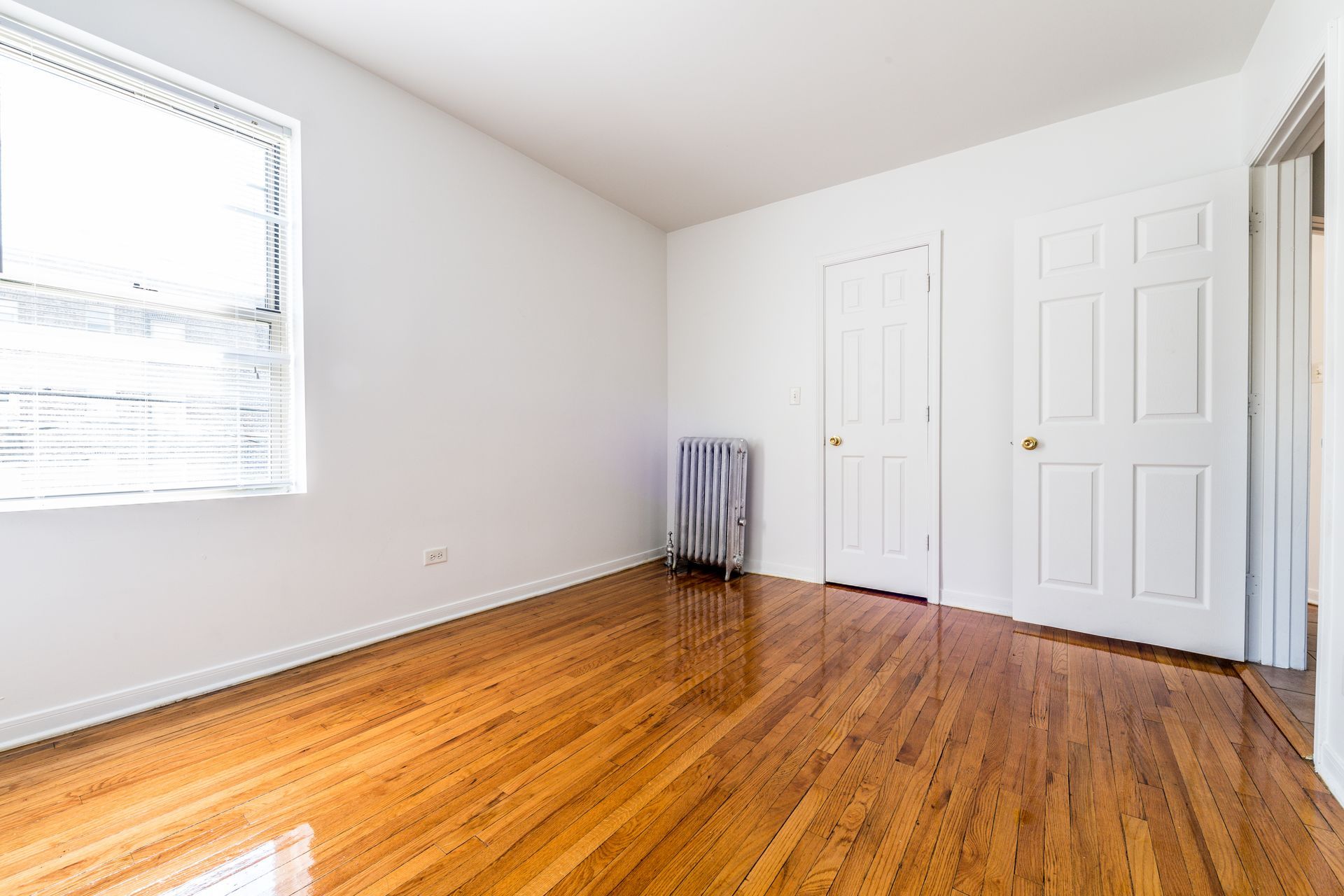 Empty room with hardwood floors, white walls, two doors, a window, and a radiator.