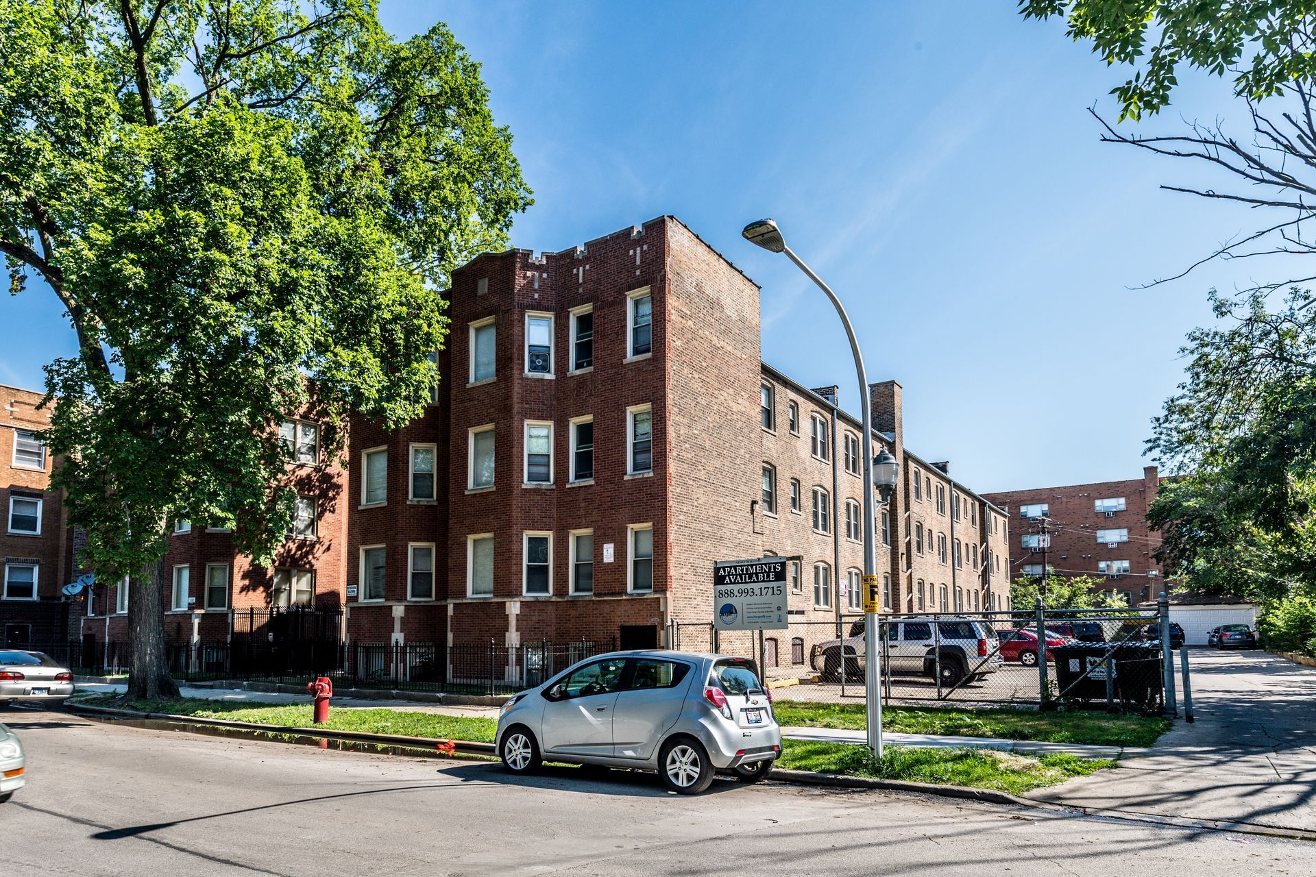 Apartment building with brick facade, parked cars, trees, and street on a sunny day.