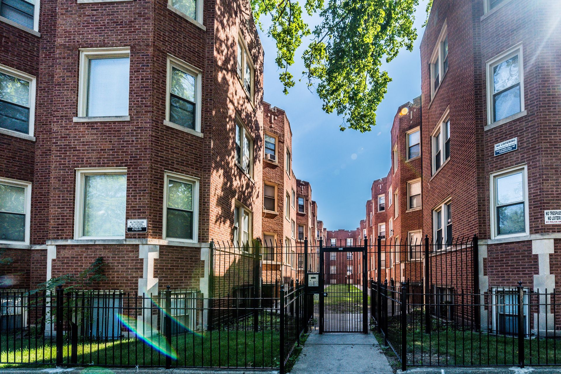 Brick apartment buildings with a gated walkway, green grass, and blue sky visible.