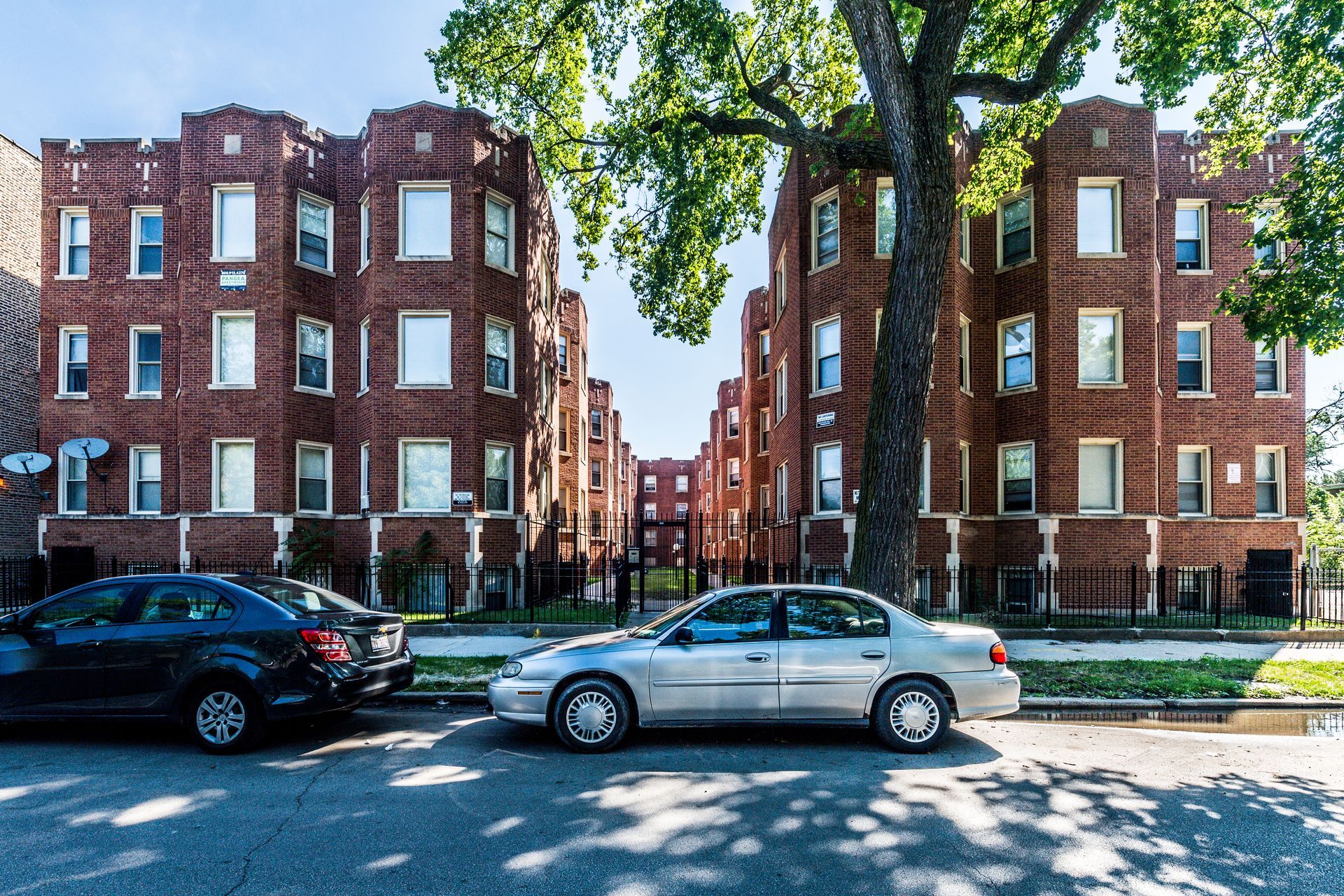 Brick apartment buildings with parked cars on street, tree in foreground.