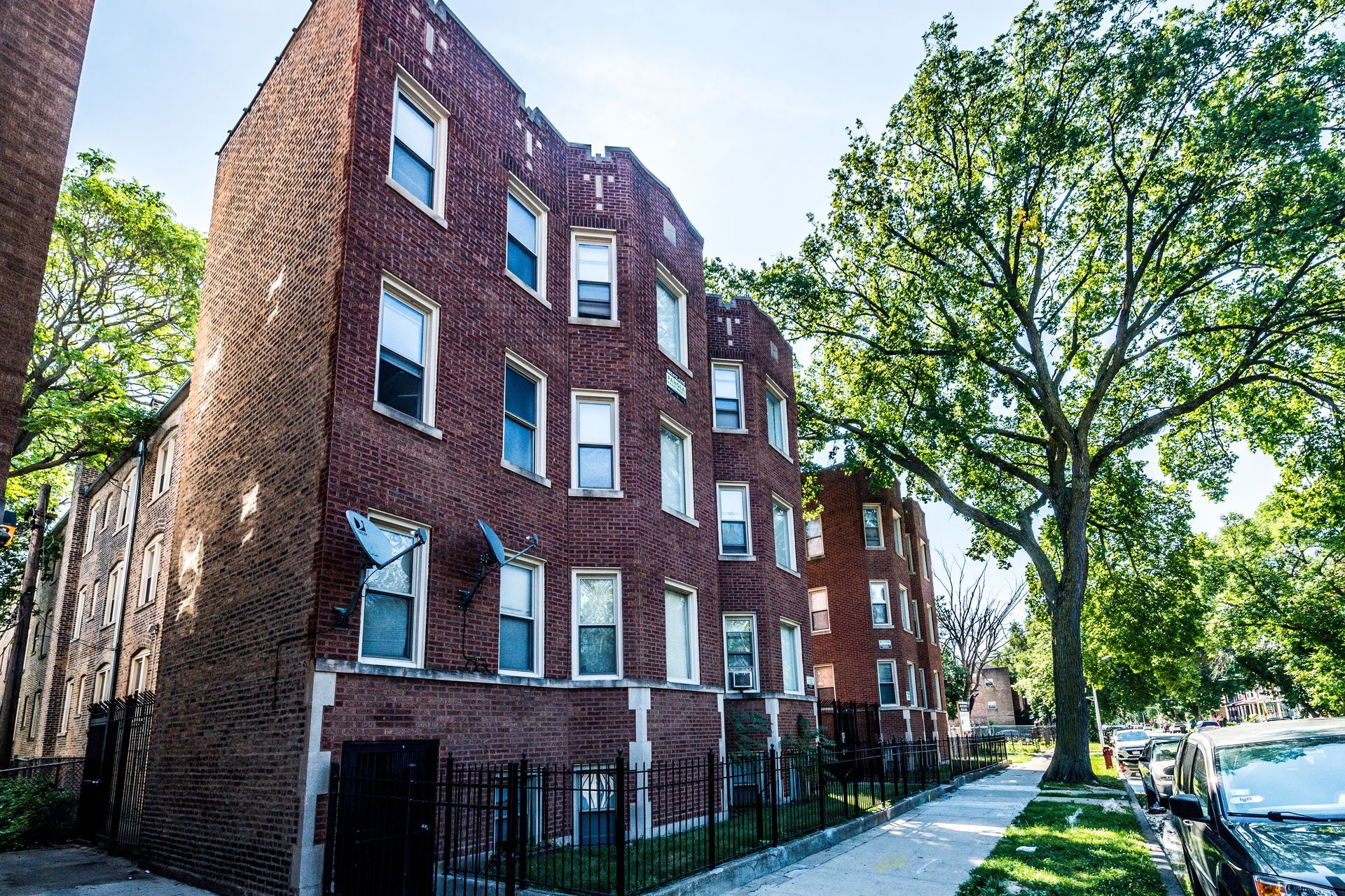 Brick apartment building with many windows, next to a sidewalk with a large tree.