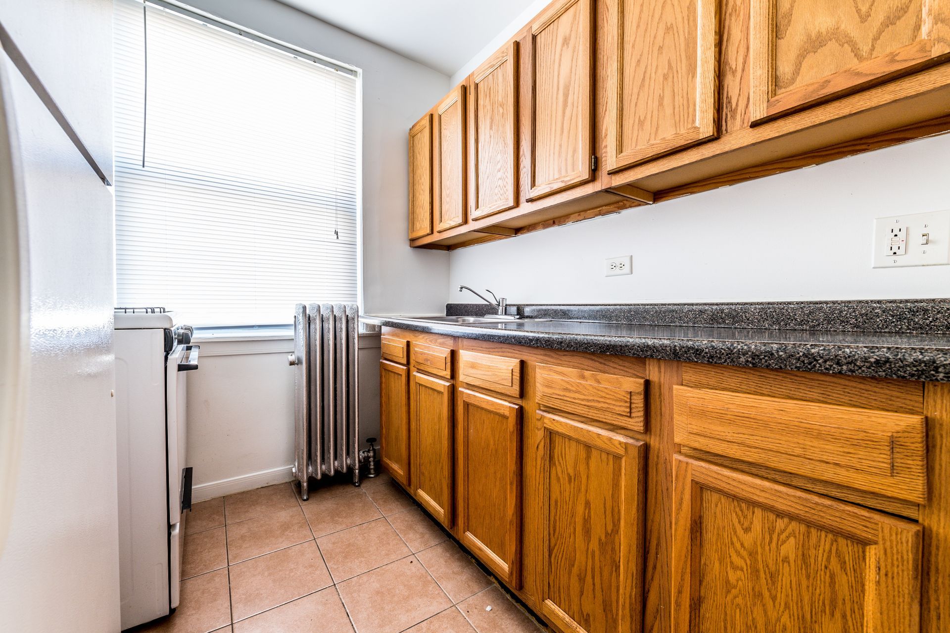 Small kitchen with wooden cabinets, countertop, sink, and window with blinds.