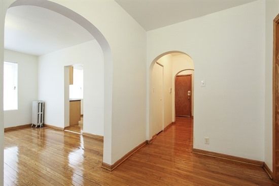 Empty apartment interior with archways, hardwood floors, and radiator.