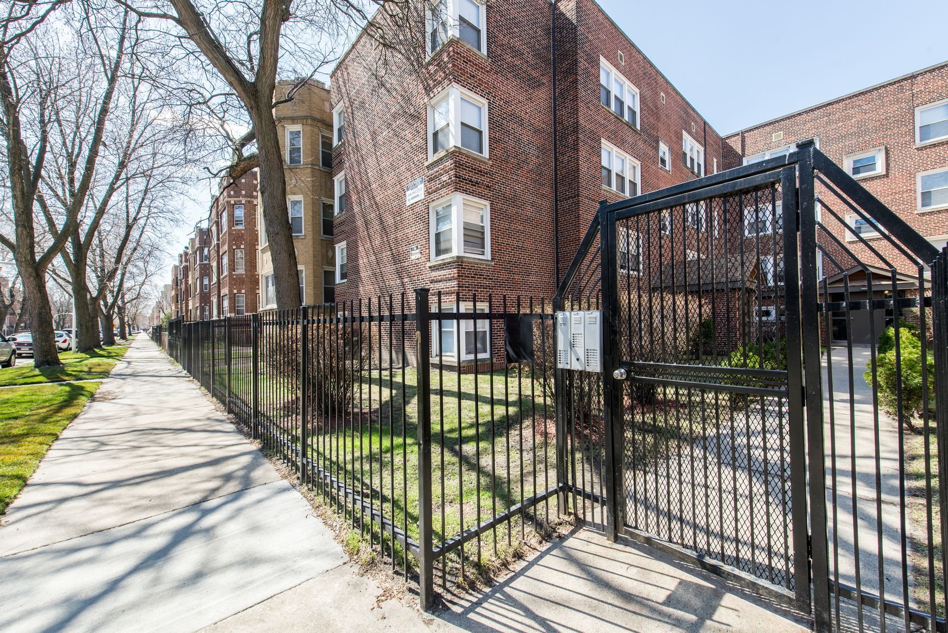 Apartment buildings with brick facade behind a black metal fence and sidewalk.