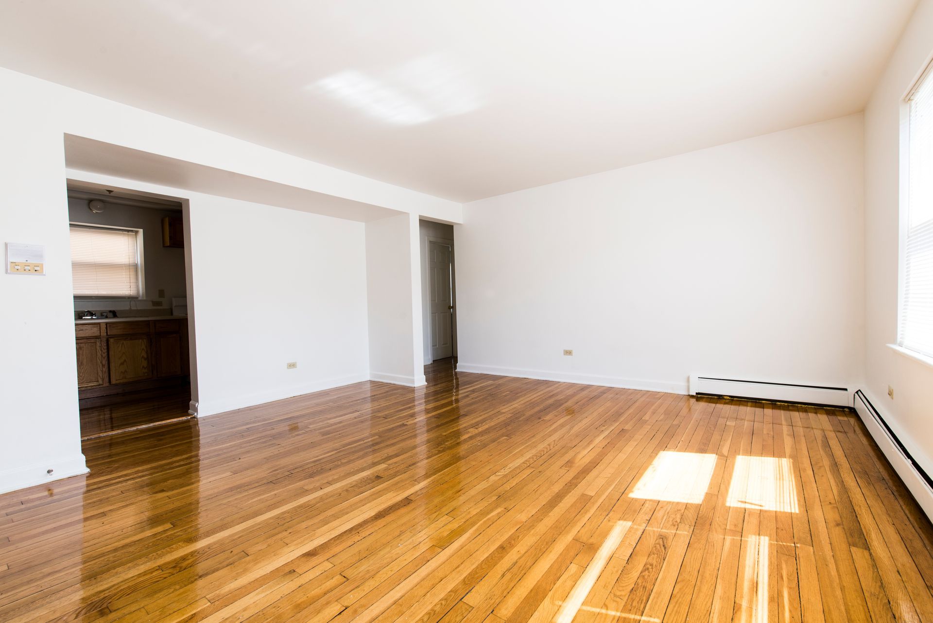 Empty room with hardwood floors, white walls, and a view into a kitchen area.