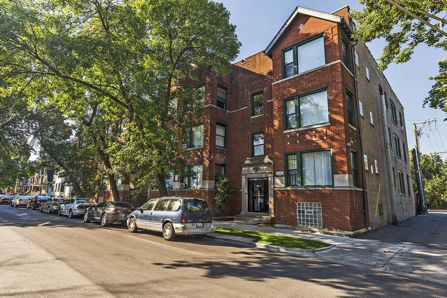 Multi-story brick apartment building on a tree-lined street with parked cars.