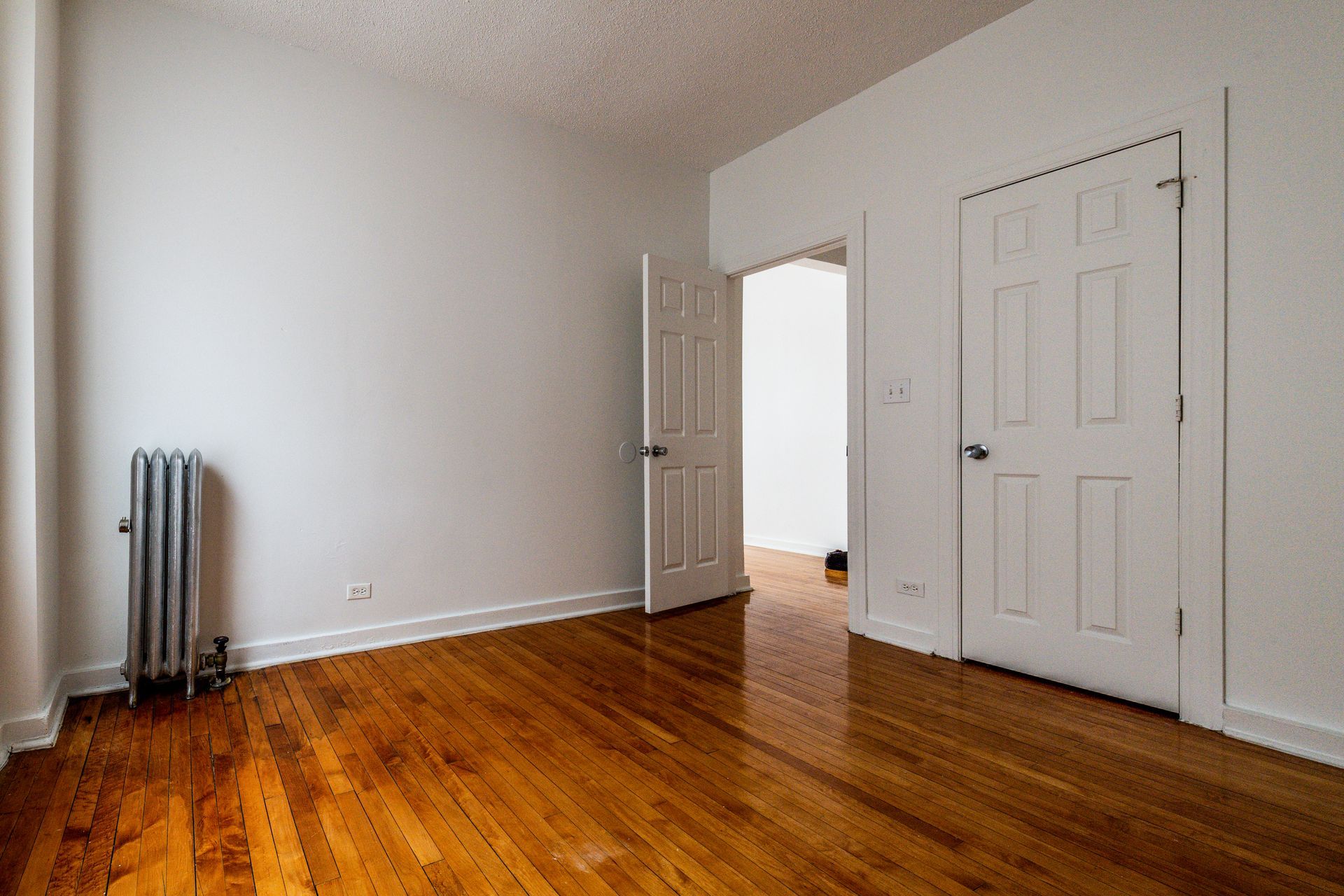 Empty room with hardwood floors, white walls, two white doors, and a radiator.