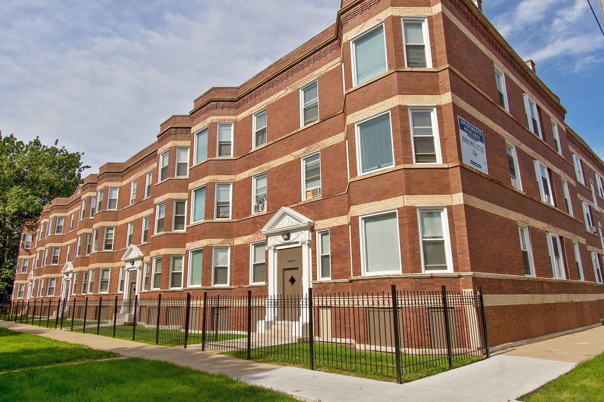 Brick apartment building with multiple windows, green lawn, and wrought iron fence.