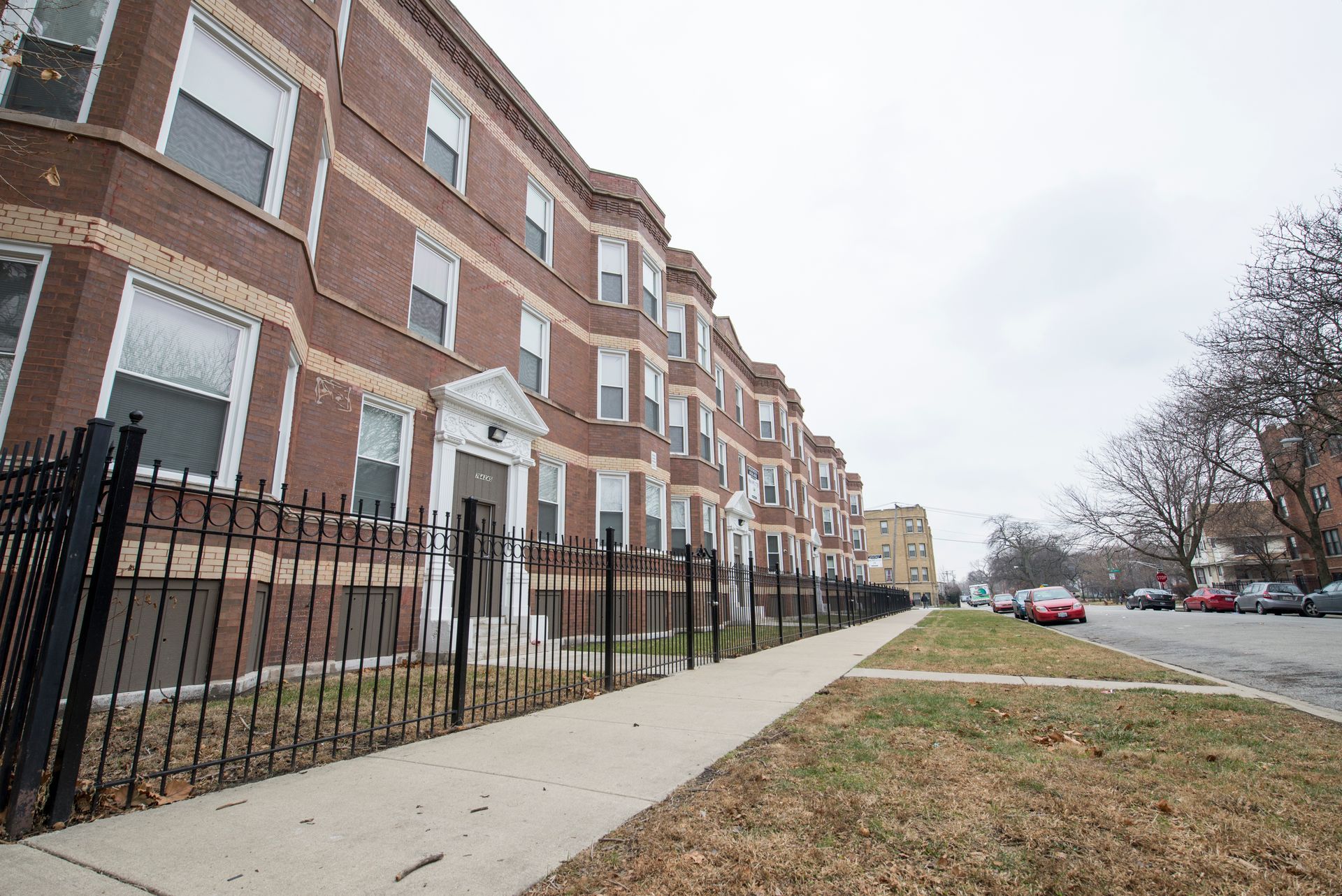 Brick apartment building with black fence, sidewalk, street, and overcast sky.