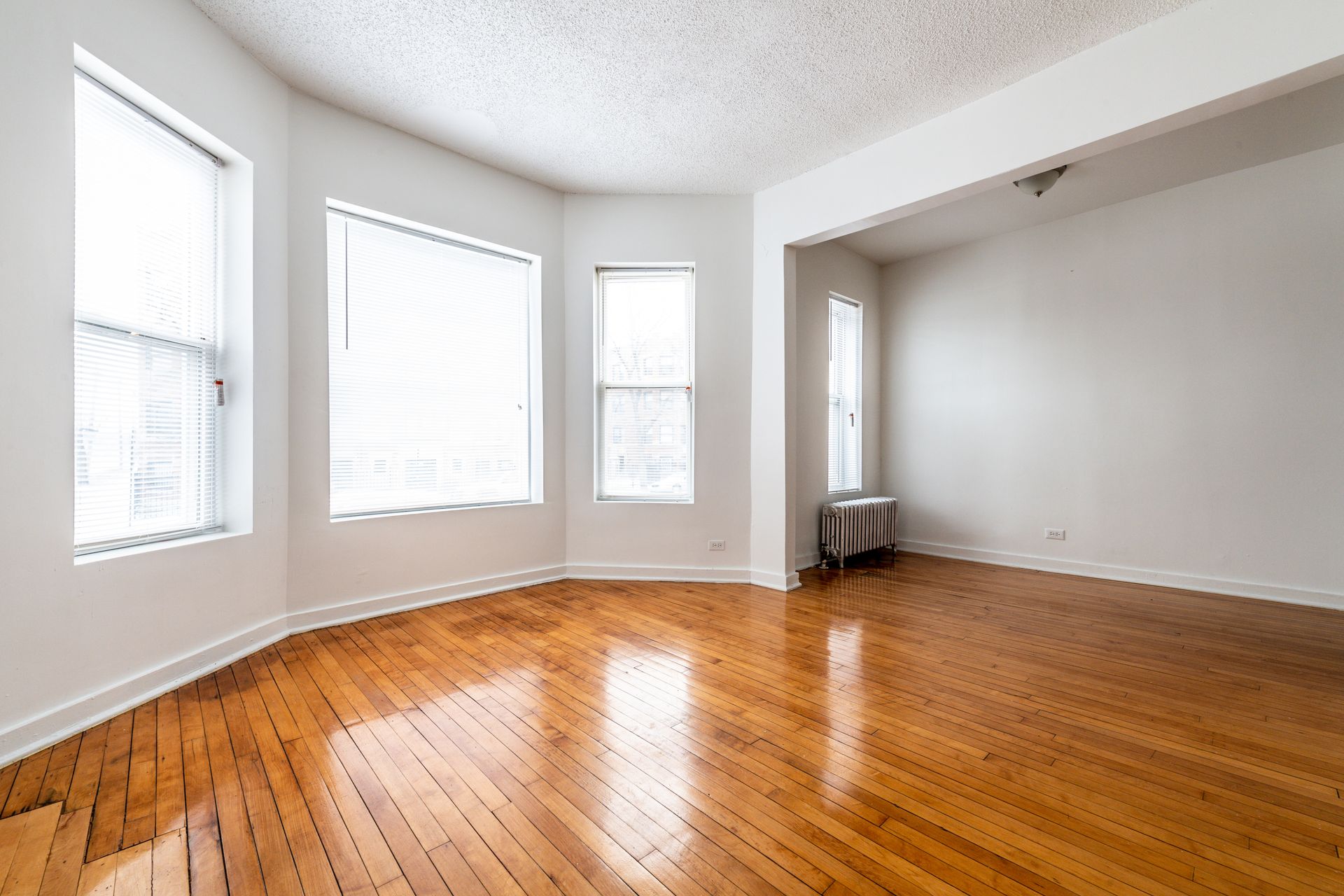 Empty room with hardwood floors, three windows with blinds, and white walls.