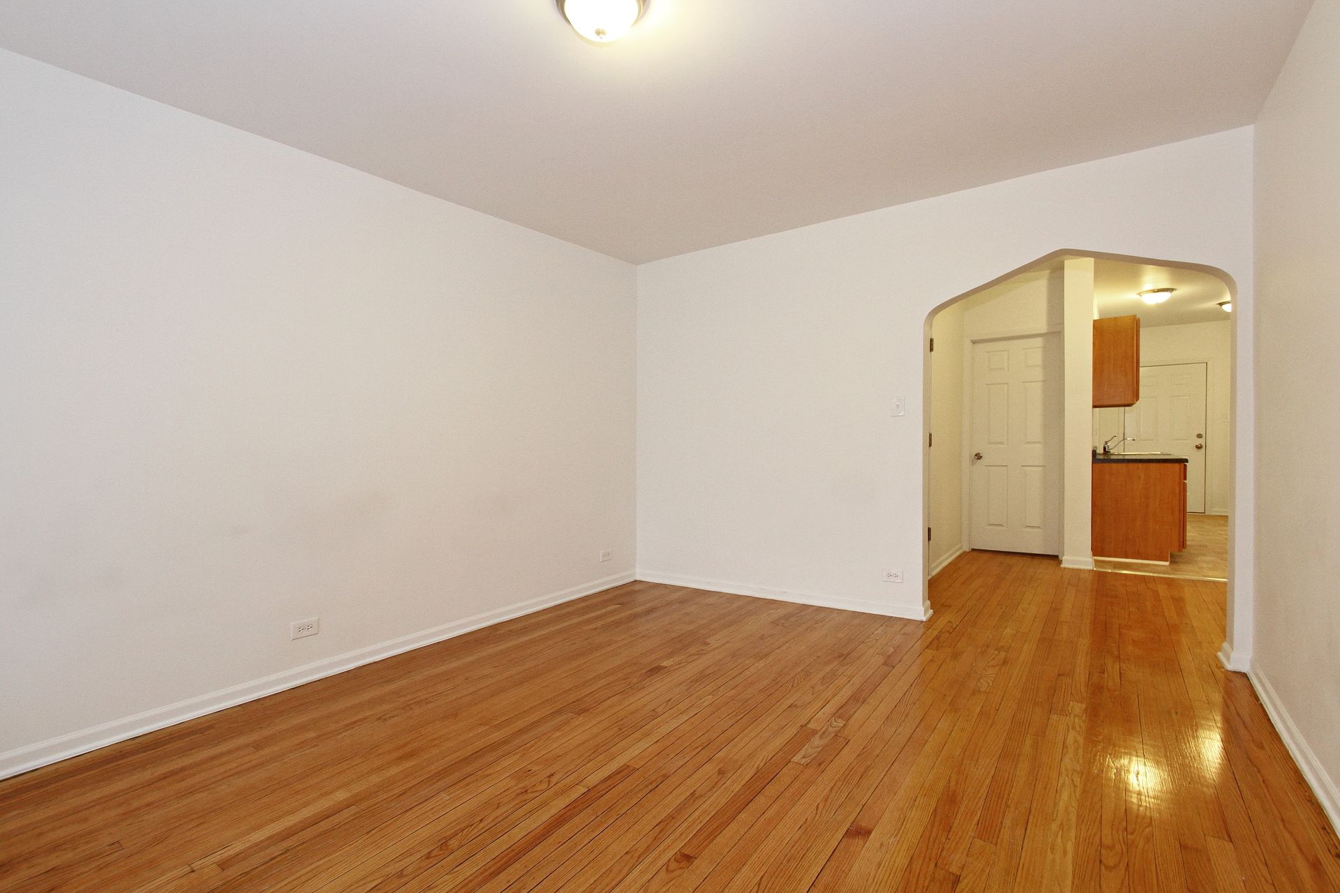 Empty room with hardwood floors, white walls, and archway leading to kitchen.