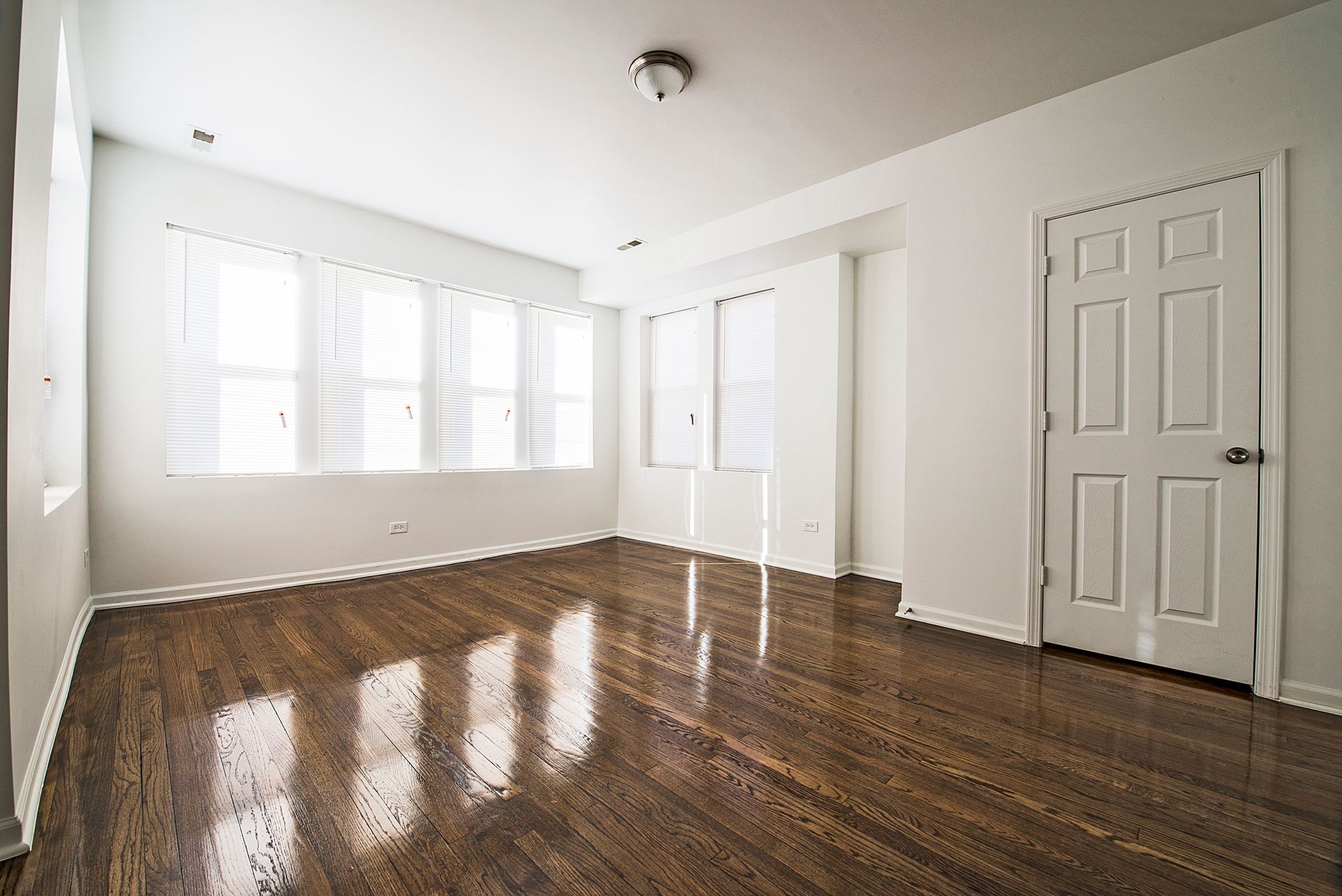 Empty room with hardwood floors, white walls, windows, and a closed door.
