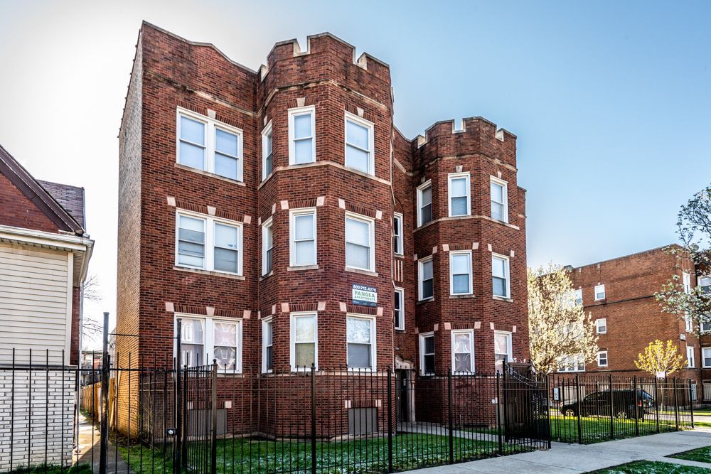 Red brick three-story apartment building with castle-like turrets and white-framed windows, behind a black metal fence.