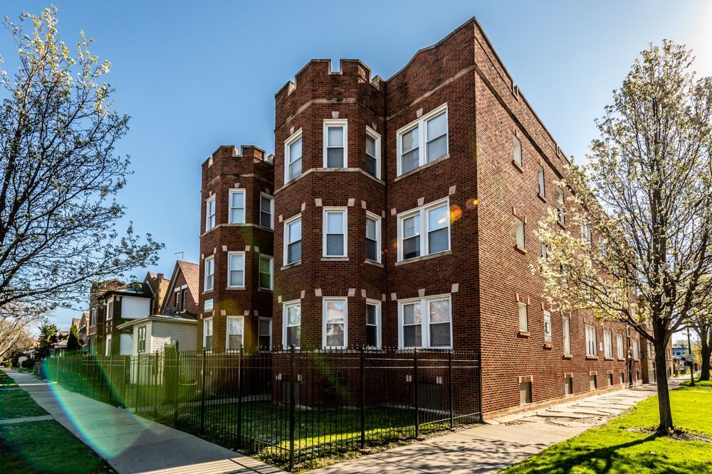 Brick apartment building with turrets on a sunny day, next to a sidewalk and green lawn.