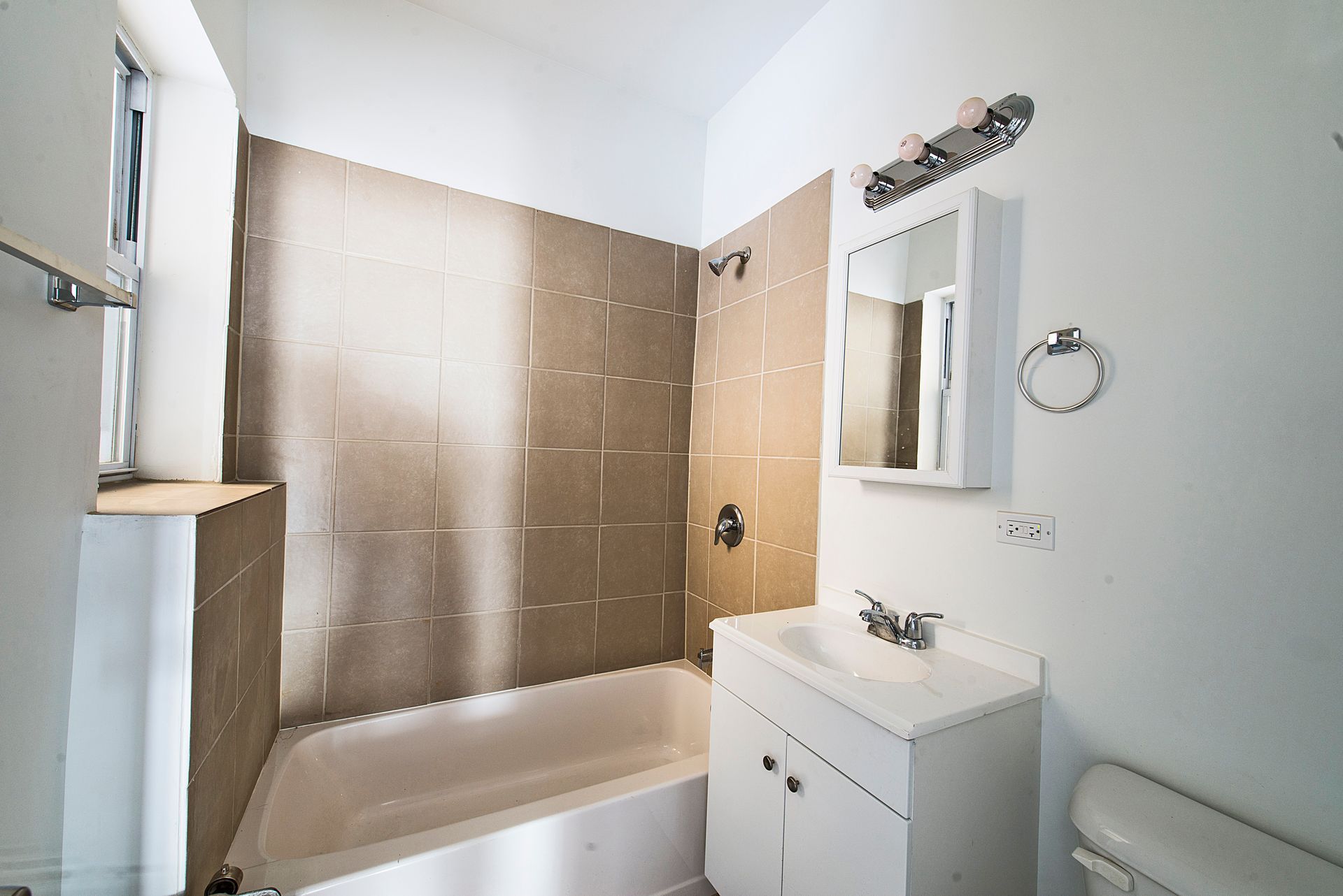 Bathroom with white walls, tan shower tiles, white sink, and toilet. Includes a small window.